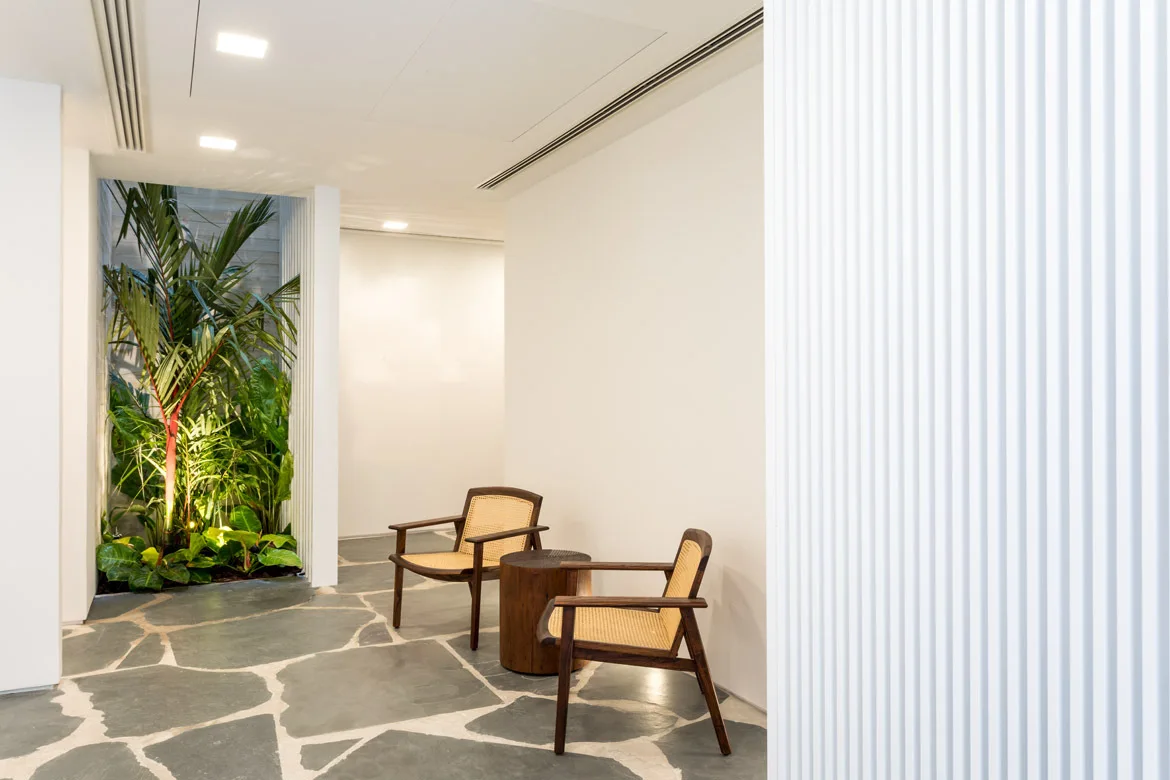 A white hallway featuring two wood and cane armchairs, a round wooden side table, irregular grey flagstone flooring, and an indoor garden alcove.