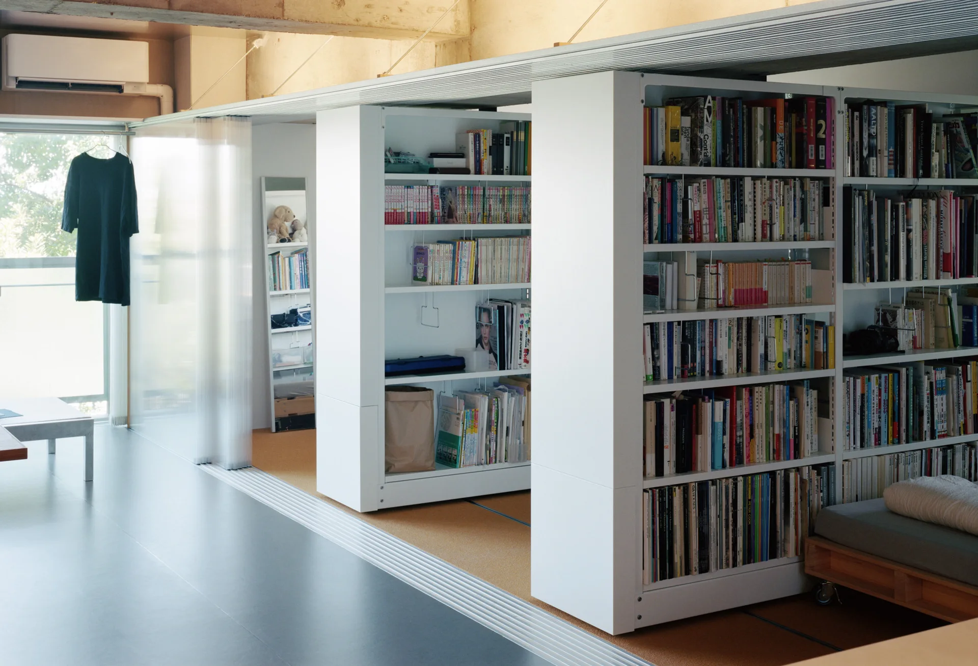Tall white bookshelves sit on a floor track system next to a translucent sliding partition beneath an exposed concrete ceiling.
