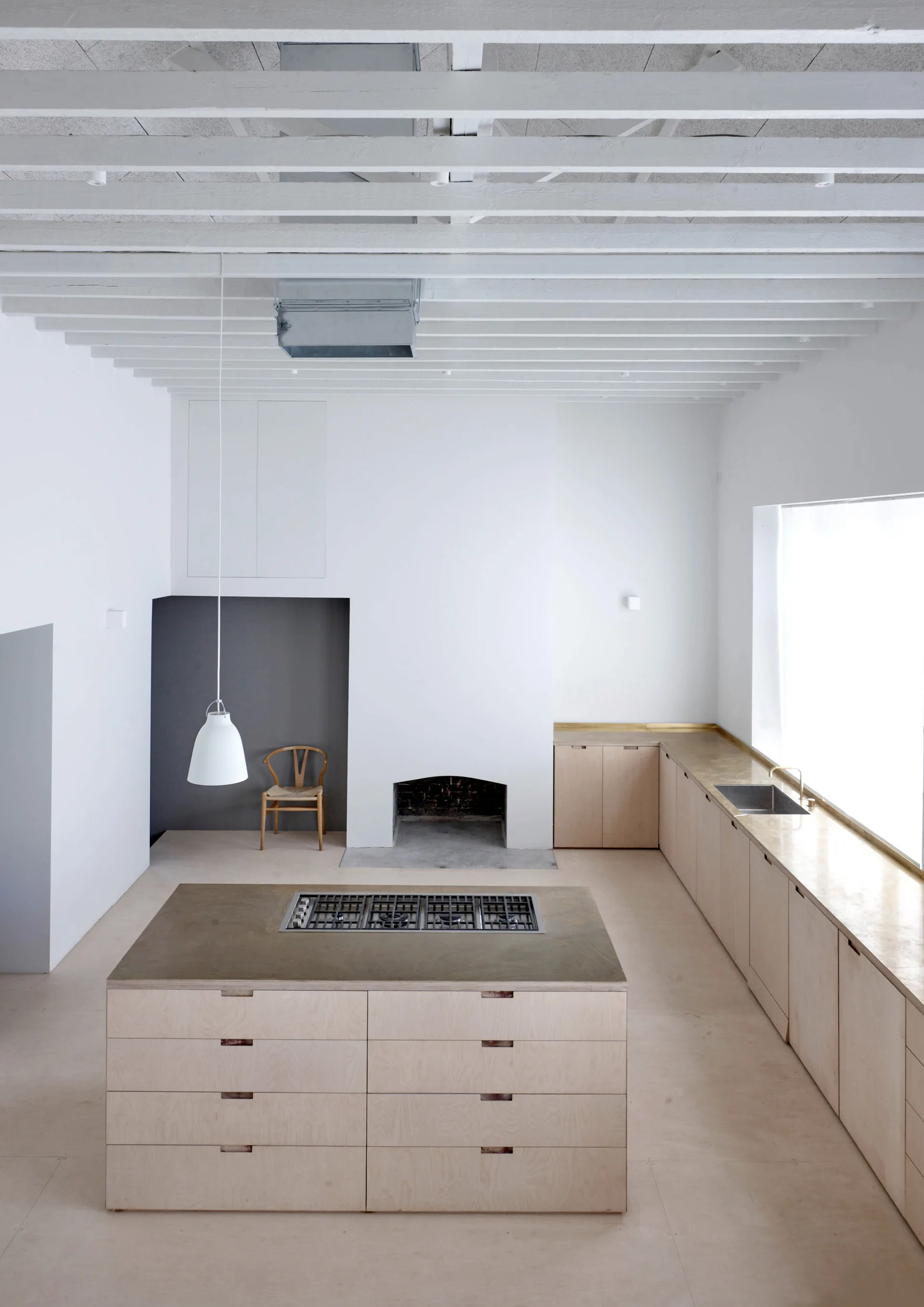 A minimalist kitchen features a light wood island, matching base cabinets, a built-in fireplace, and painted white ceiling beams.