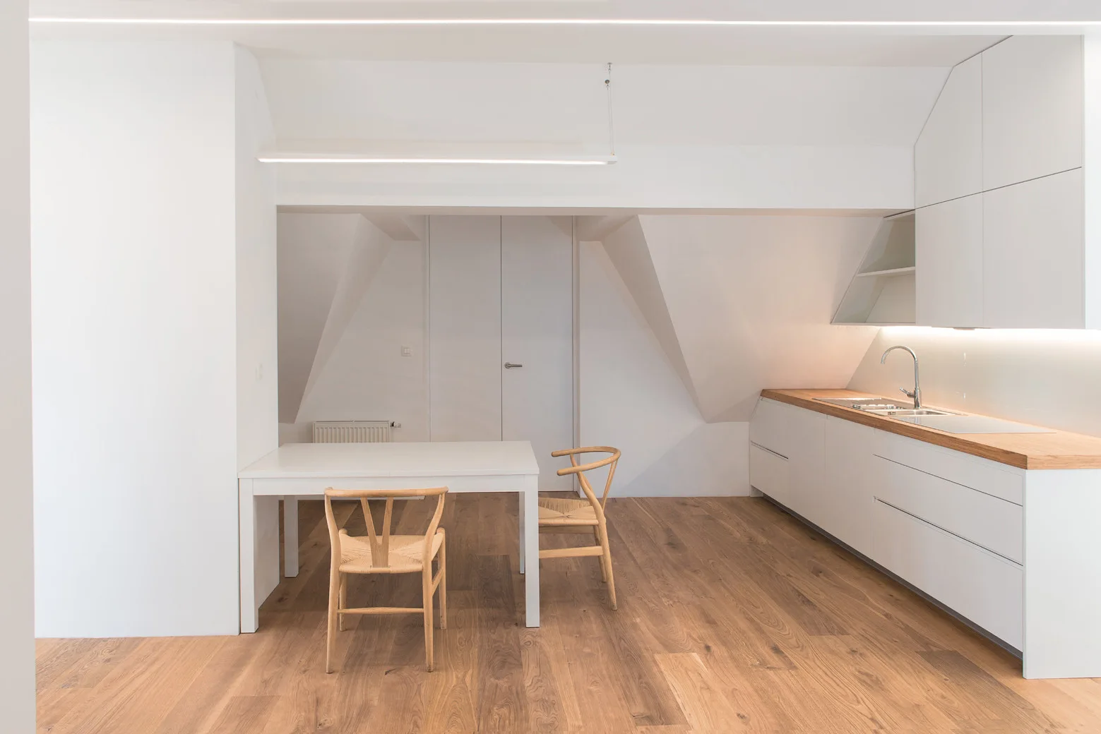 A minimalist white attic kitchen features flat-front cabinetry with wood countertops, a white dining table, and wooden chairs.