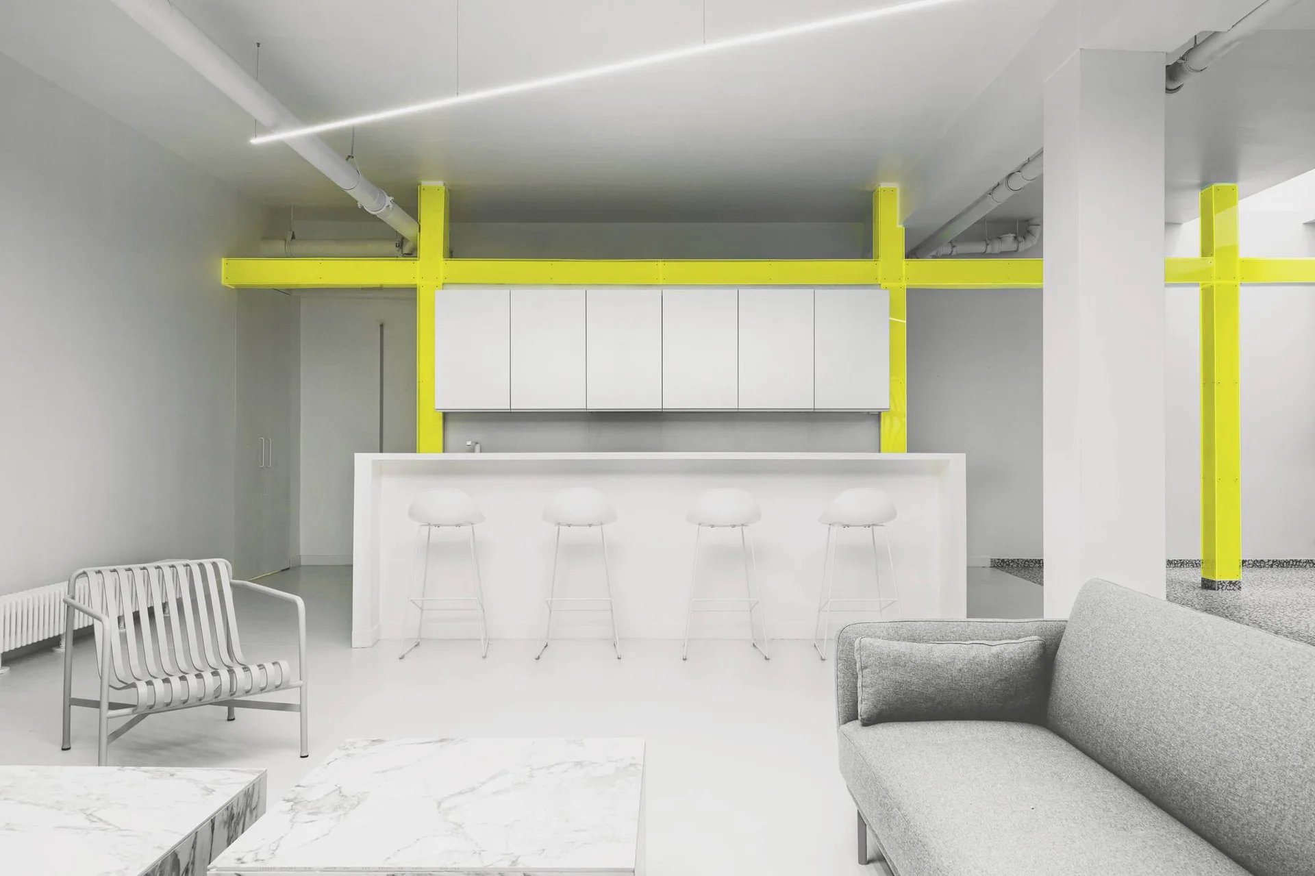 A minimalist open-plan kitchen and living area featuring white cabinetry, a long white island with stools, and vibrant yellow structural steel beams.