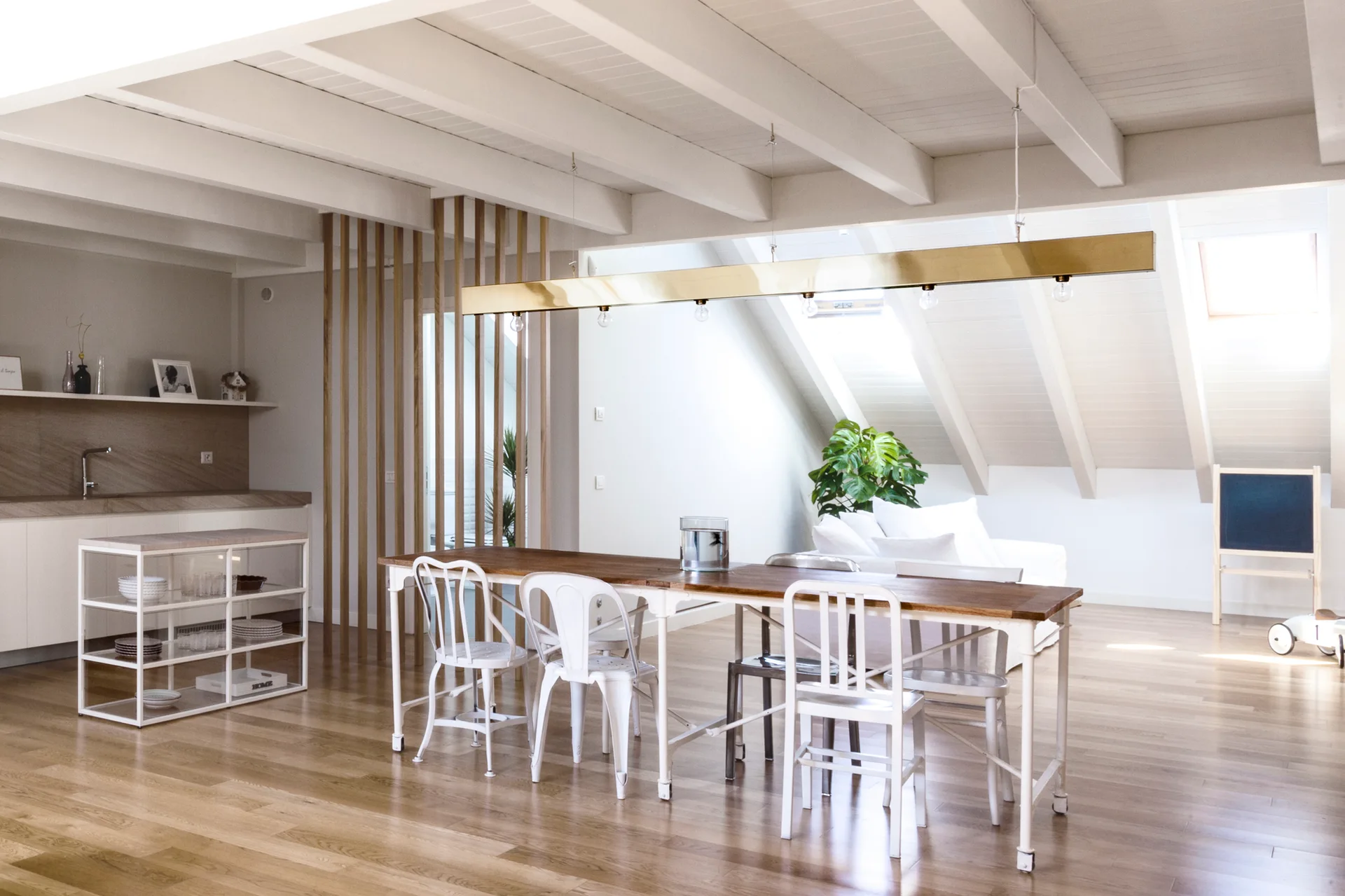 A light-filled attic dining room features a long wooden table with white metal chairs under a brass pendant light and white exposed ceiling beams.