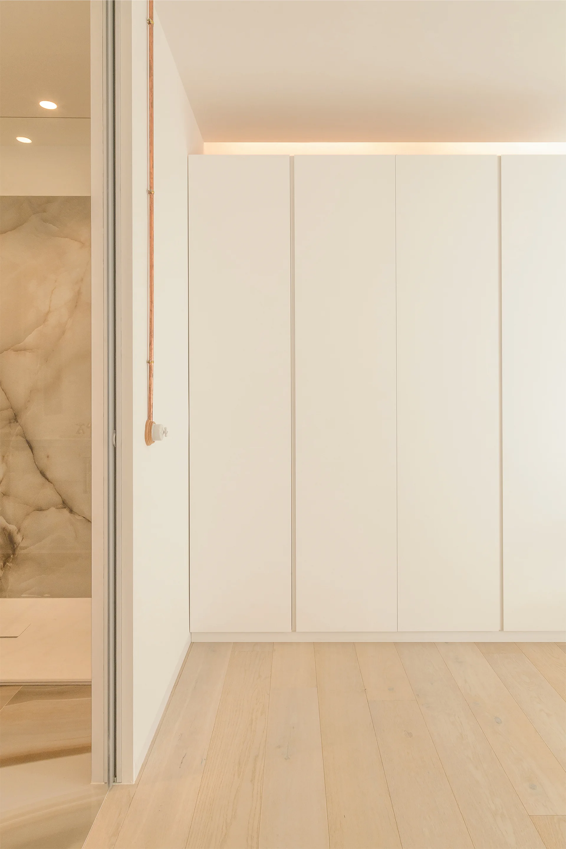 A minimalist dressing area features seamless white wardrobe cabinets, light wood flooring, and an exposed copper pipe on a white wall.