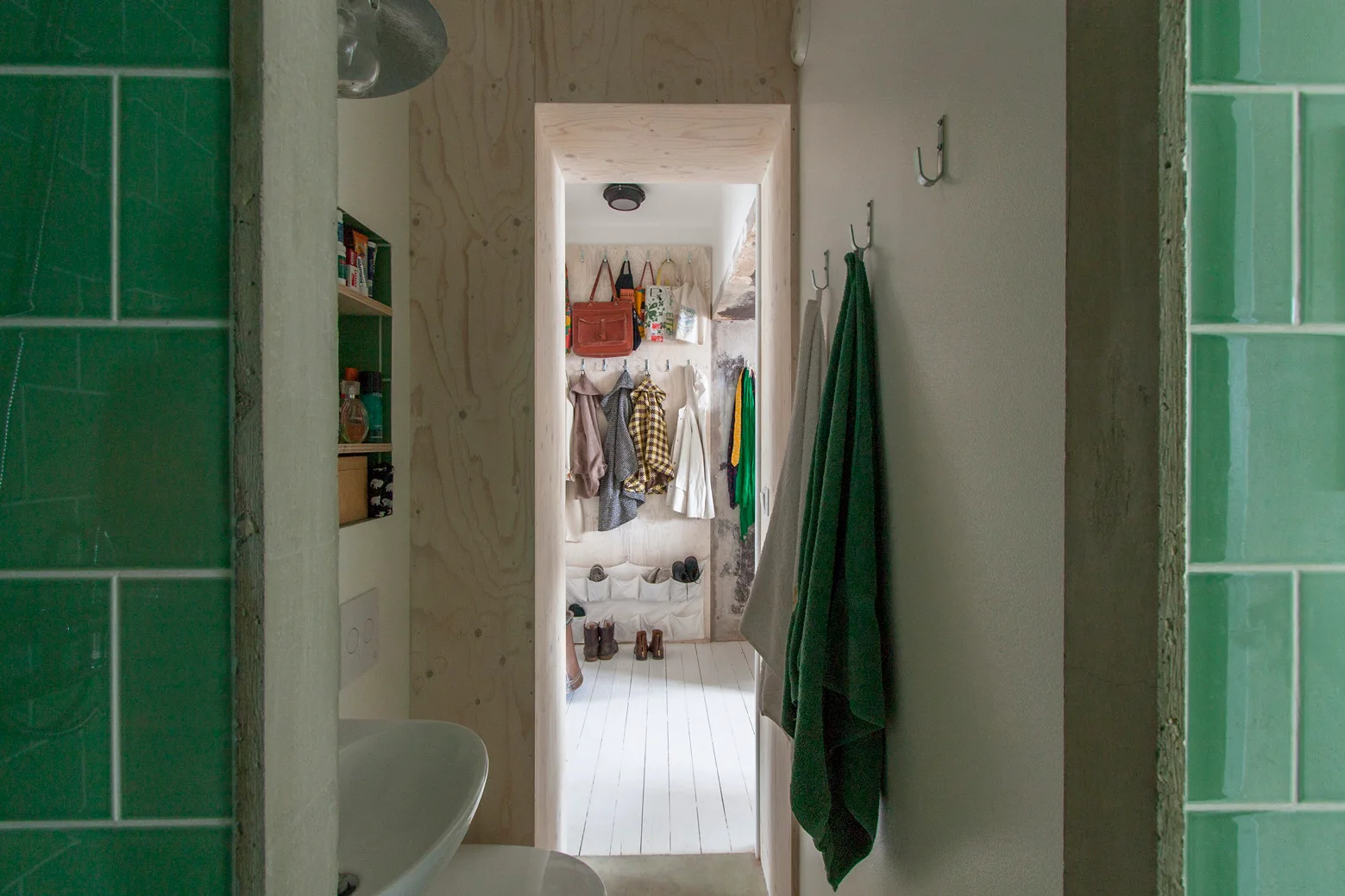 A view through a raw plywood doorway from a green-tiled bathroom looking into a bright mudroom with coat hooks and white floorboards.