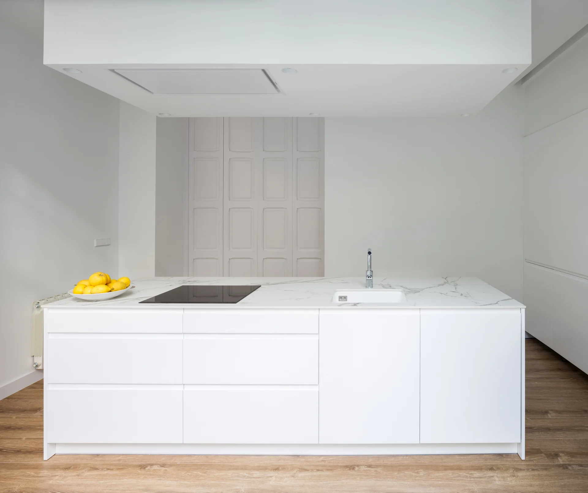 A minimalist white kitchen island features a veined marble countertop, integrated sink, and black stovetop over light wood floors.