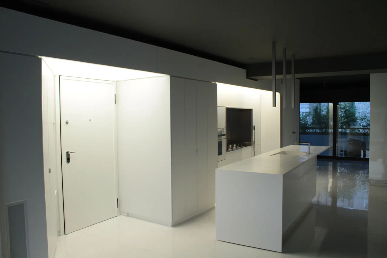 A minimalist white kitchen features a solid kitchen island, integrated handleless cabinetry illuminated by cove lighting, and a dark ceiling.