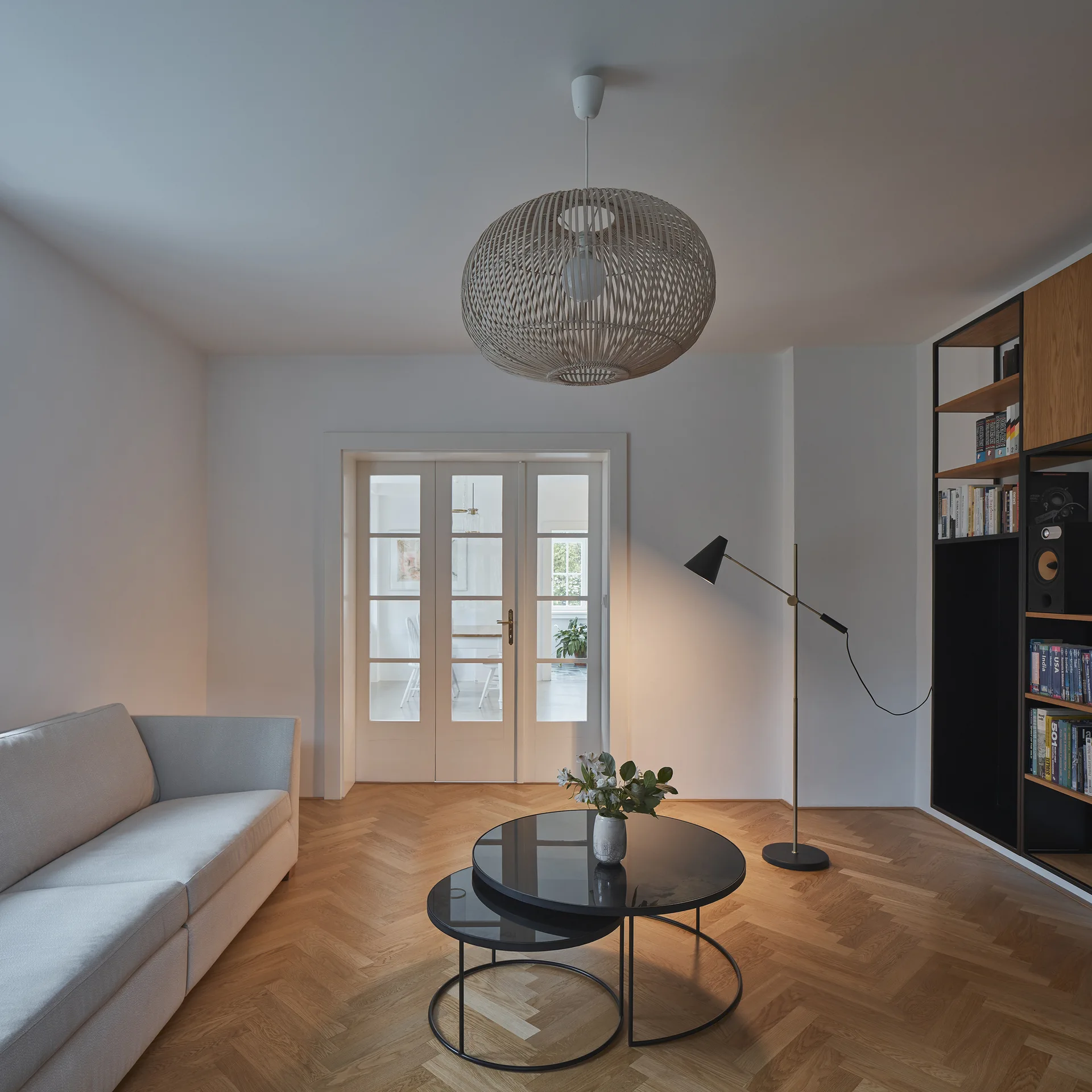 A minimalist living room features a grey sofa, black nested coffee tables, a large wicker pendant light, and white French doors.