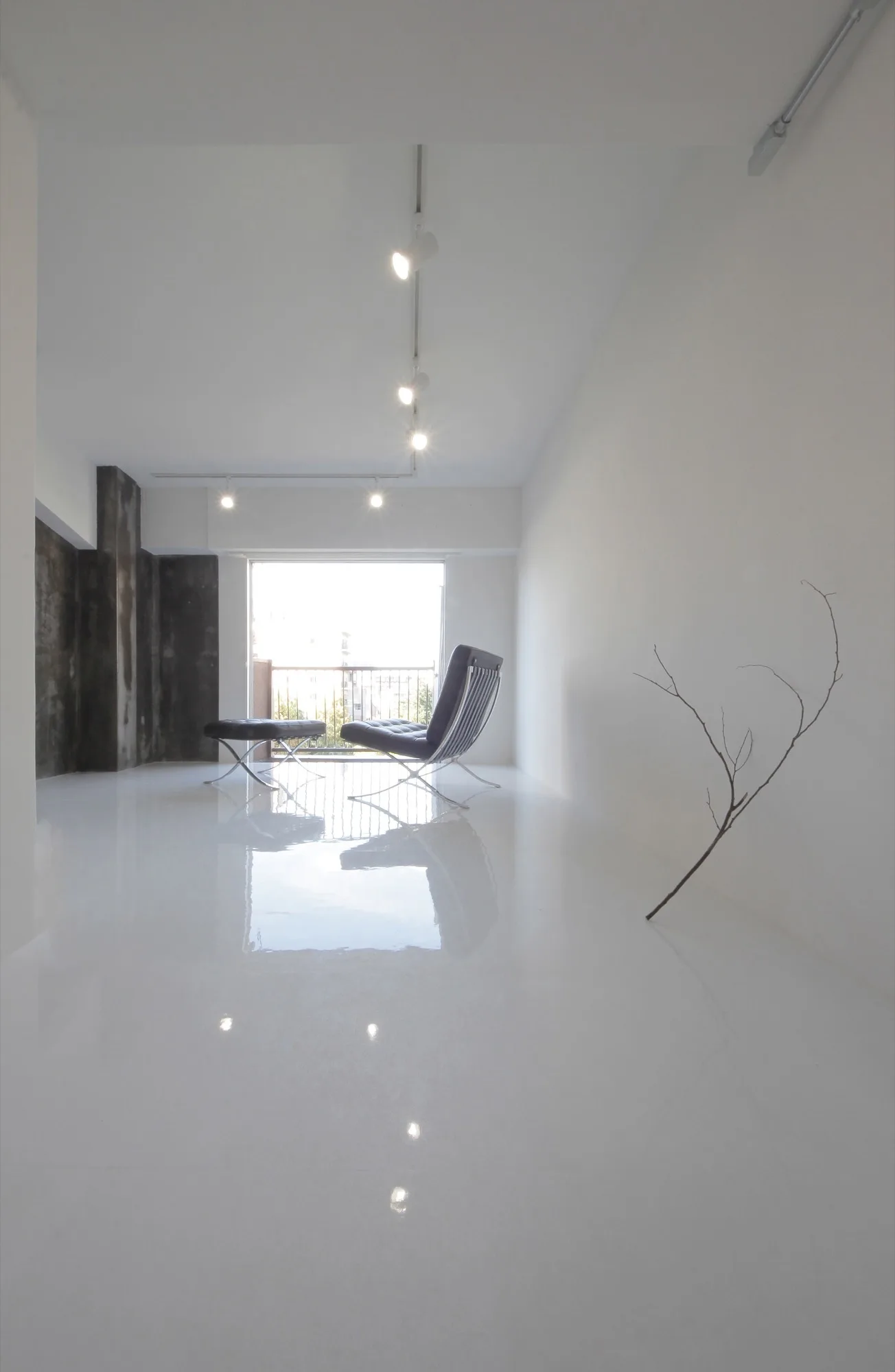A minimalist white living room features a black leather lounge chair on a highly reflective high-gloss floor with track lighting and a bare branch.
