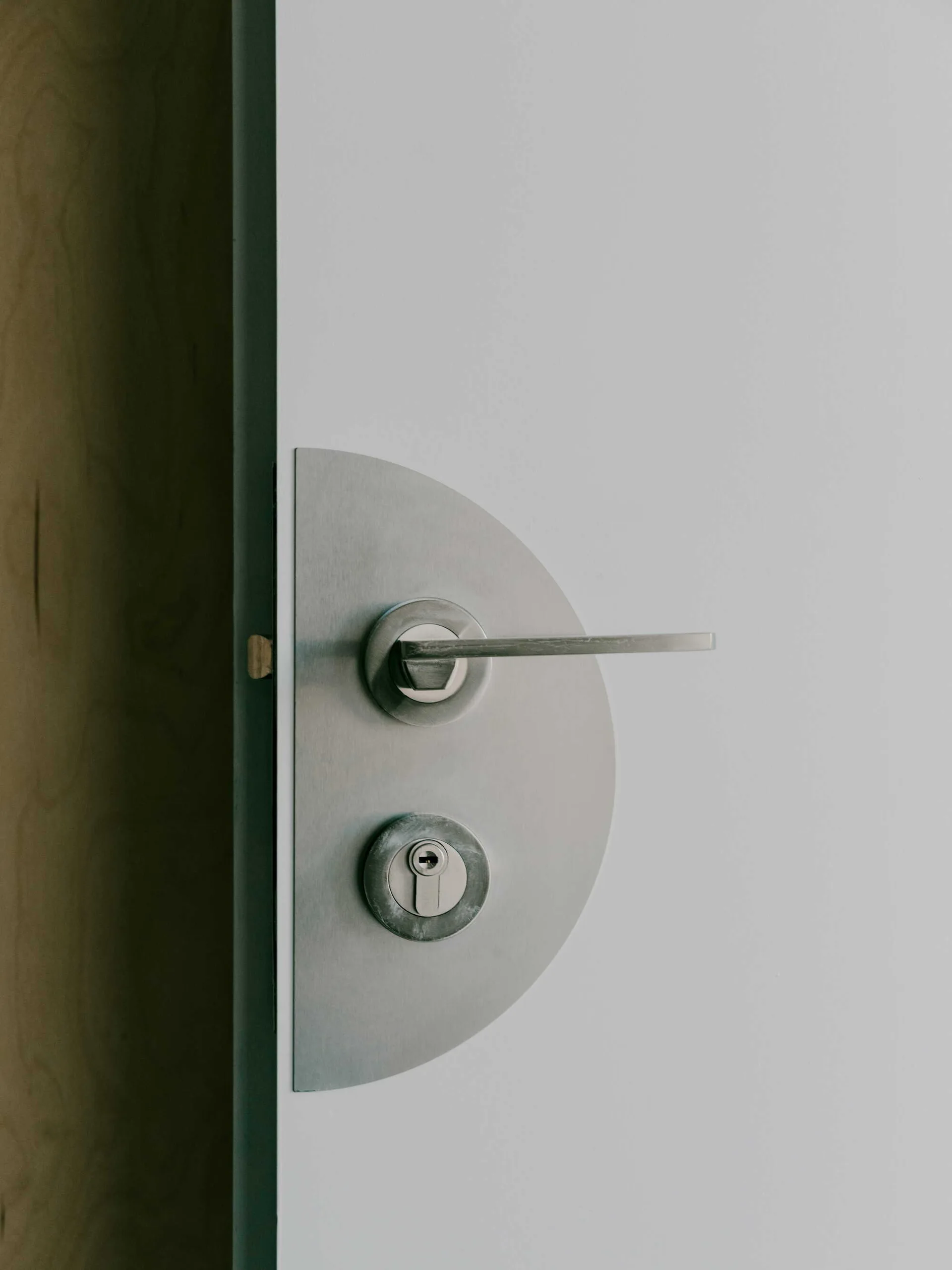A close-up of a minimalist white door featuring a semi-circular brushed metal backplate, a lever handle, and a keyed lock cylinder.