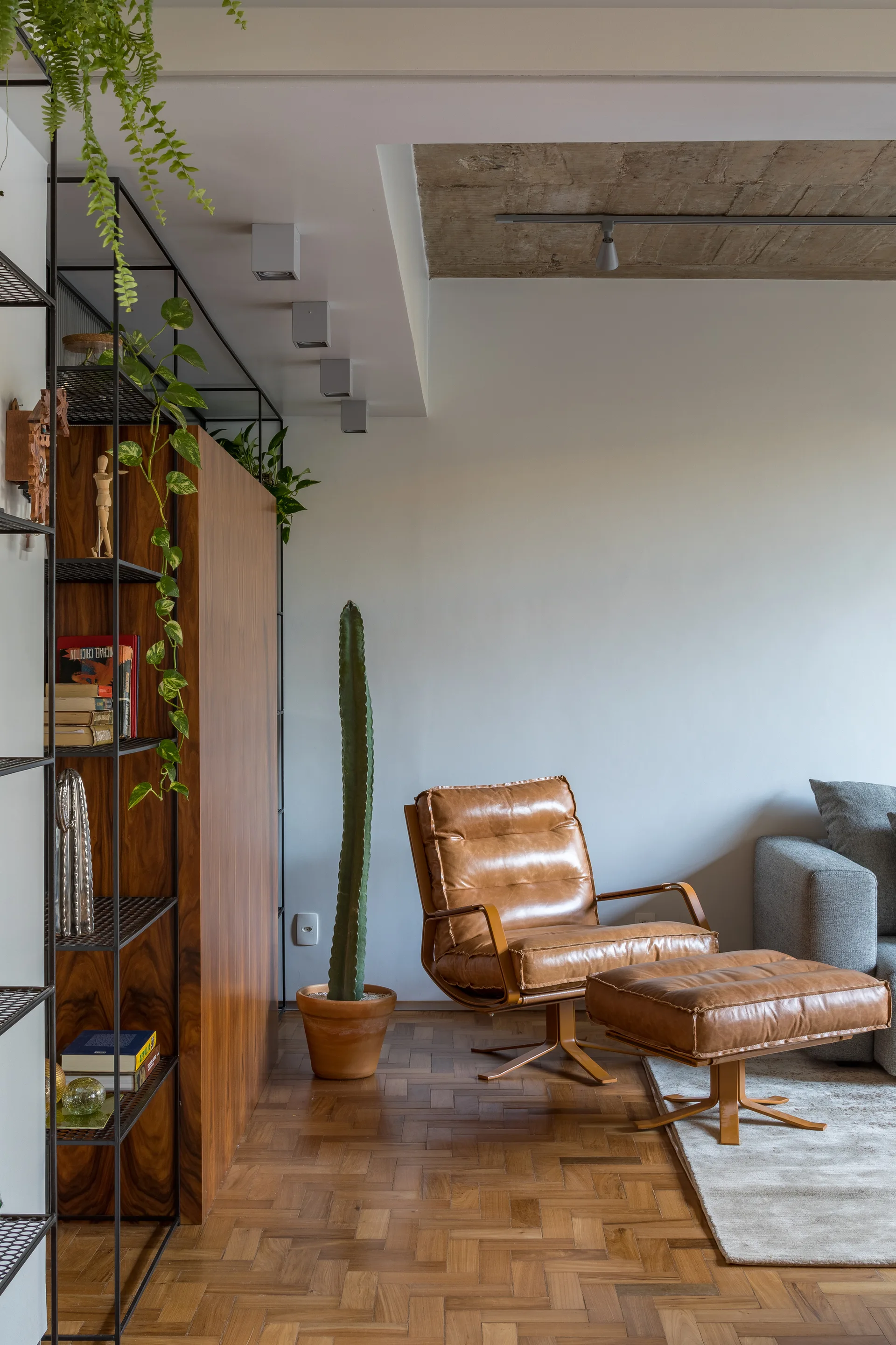 A living room corner features a tan leather lounge chair with ottoman, a tall columnar cactus, and a wooden shelving unit on herringbone parquet flooring.