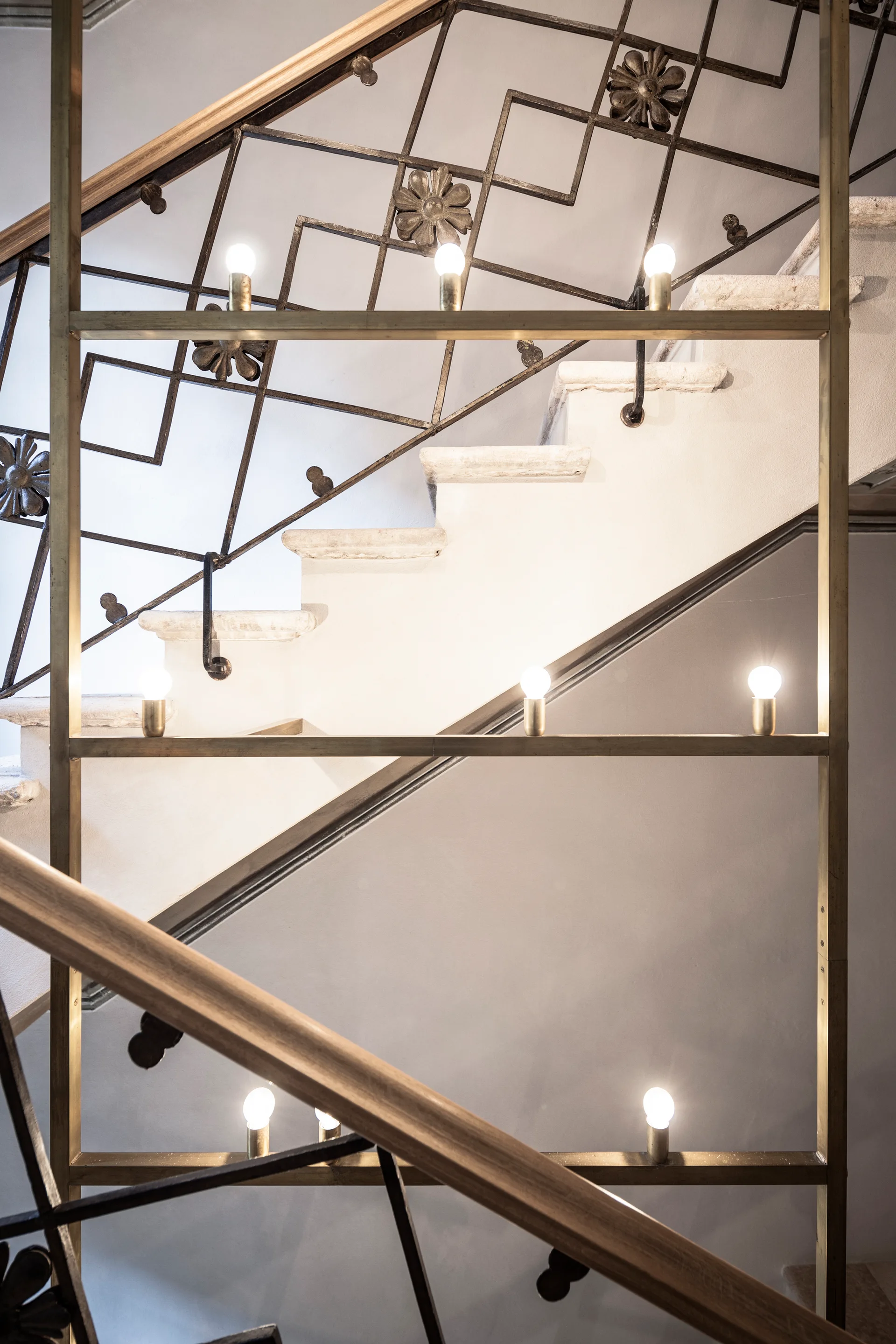 A stone staircase features geometric black iron railings with floral details next to a vertical brass frame with glowing exposed light bulbs.