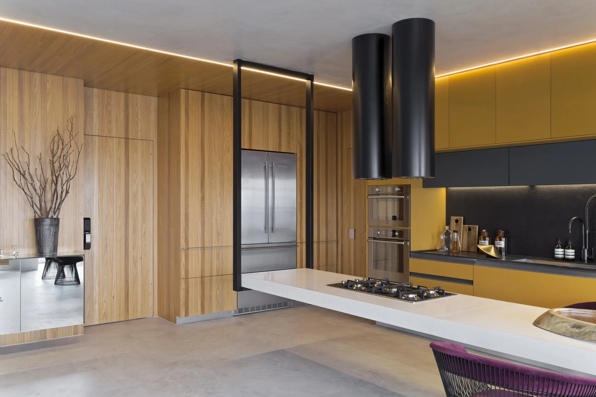 A modern minimalist kitchen featuring mustard yellow cabinetry, natural wood wall paneling, a white island, and dual cylindrical black range hoods.