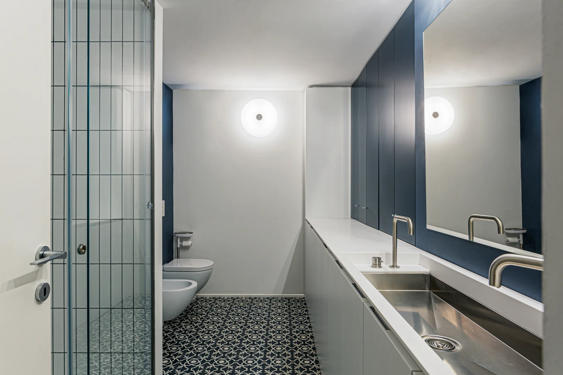 A narrow modern bathroom features a long white vanity with a metallic sink, navy cabinets, wall-mounted fixtures, and geometric patterned floor tiles.
