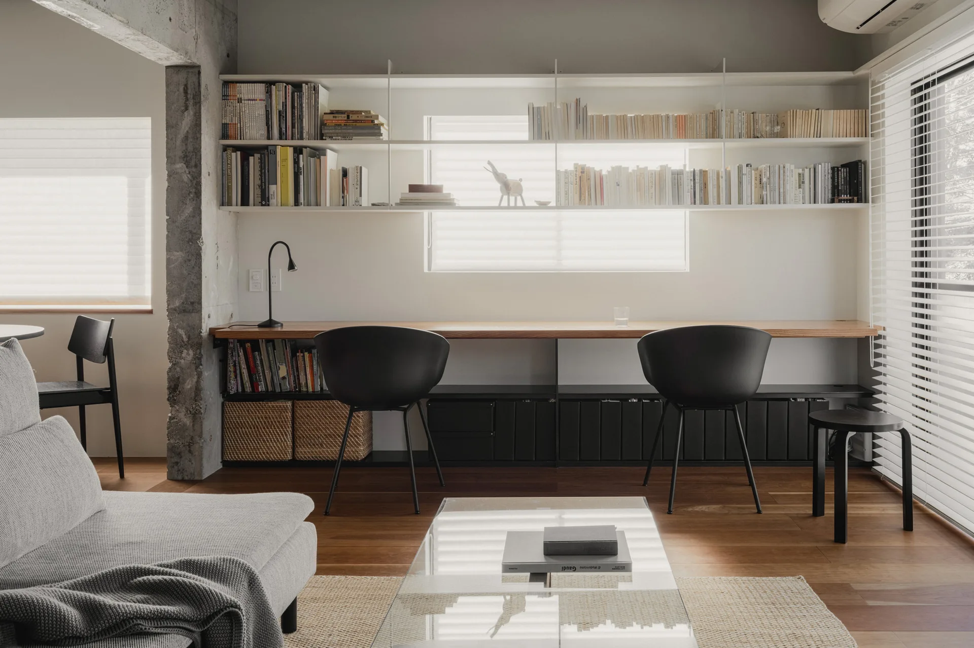 A minimalist home office featuring a long wooden desk with black chairs, white wall-mounted bookshelves, and an exposed concrete column.