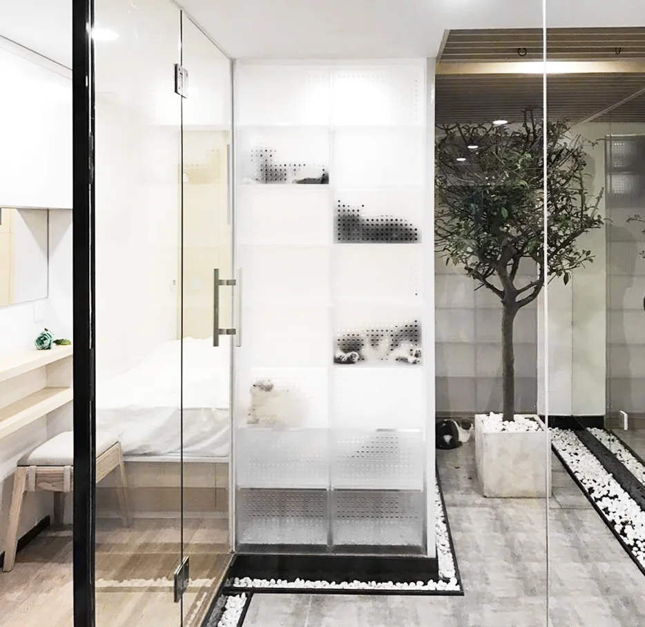 A modern interior space featuring a transparent glass partition wall, a white perforated modular cat enclosure, and a potted tree with pebble borders.