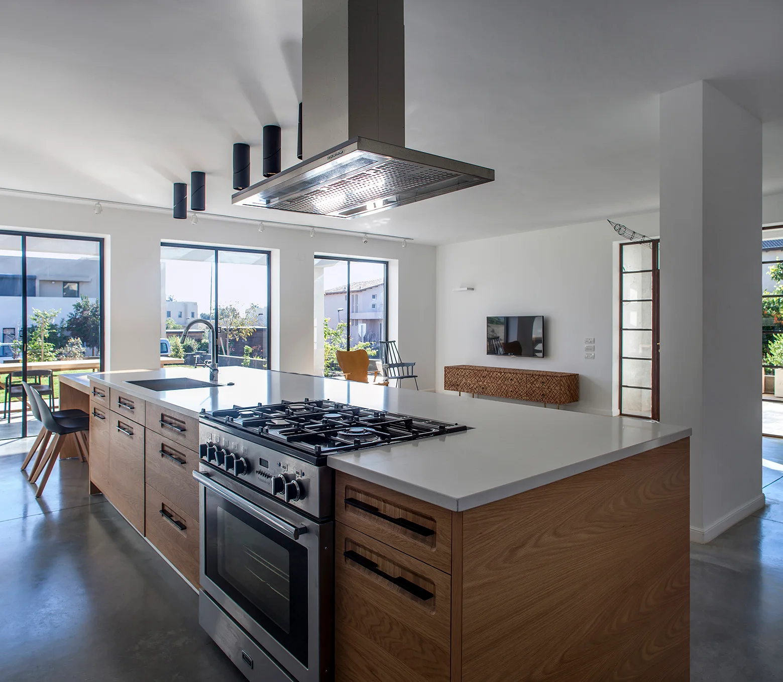 A modern kitchen featuring a large wood-framed island with a white countertop, gas range, ceiling-hung stainless steel hood, and polished concrete floors.