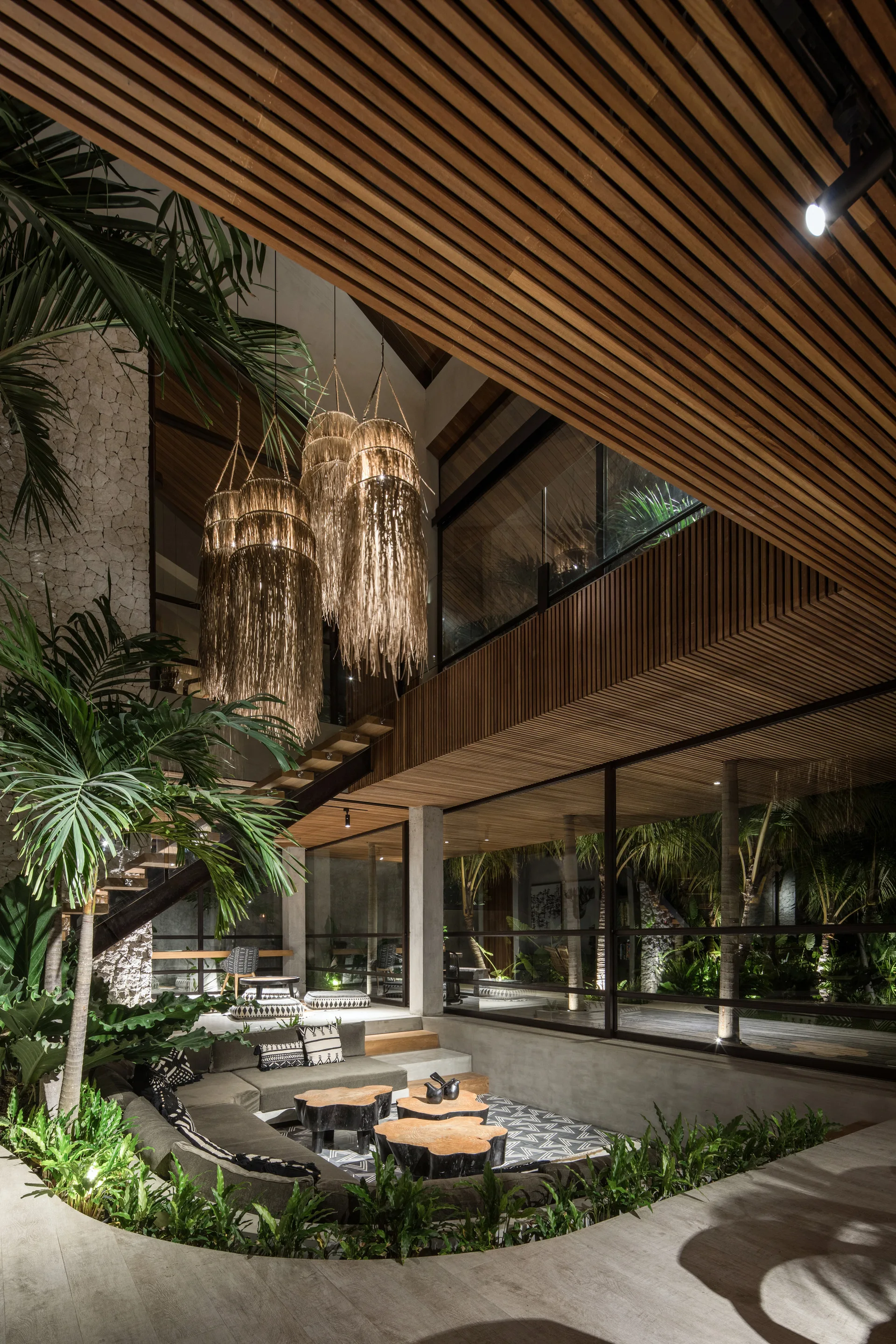 A sunken tropical living room features a curved grey sectional sofa, organic wood coffee tables, fringed pendant lights, and integrated indoor greenery.