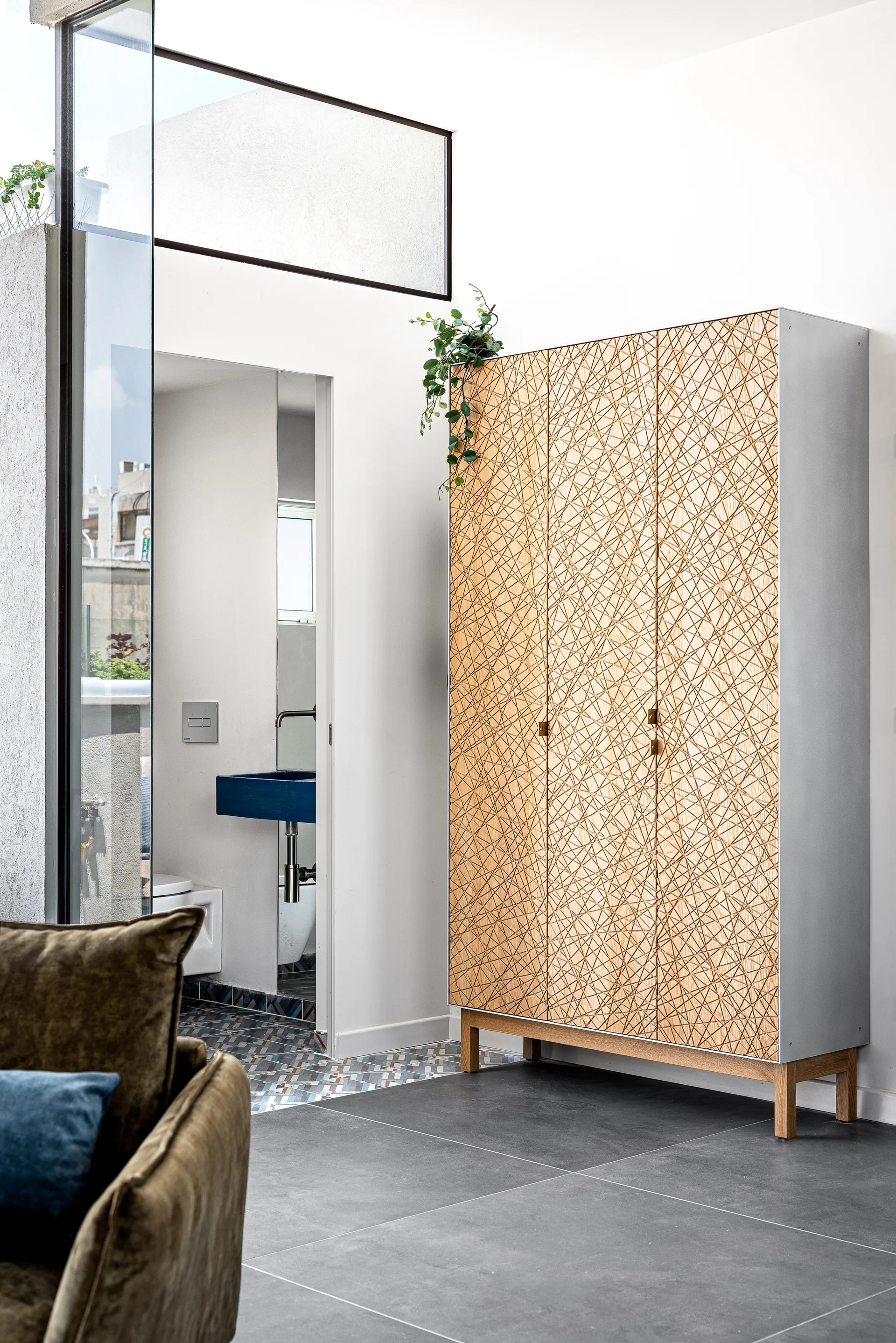 A tall modern wardrobe with geometric wood-inlay doors stands on a grey tiled floor next to an open doorway showing a blue bathroom sink.