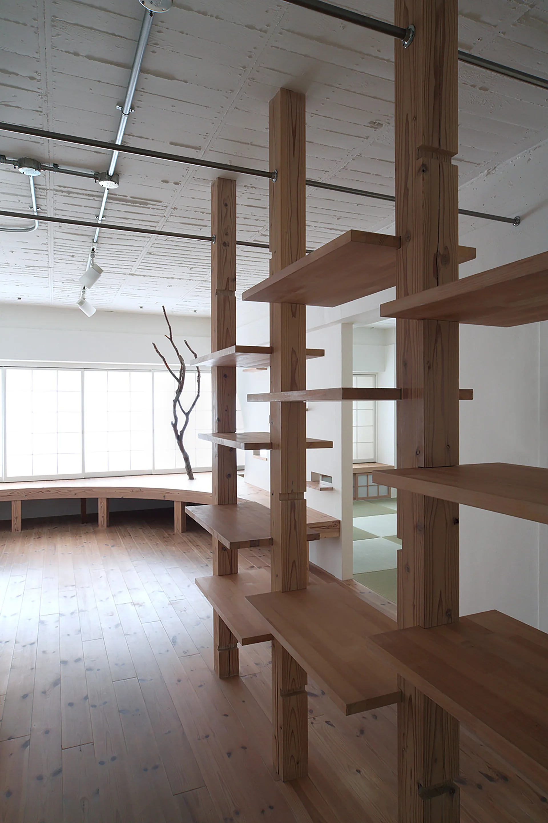 An open interior featuring a tall light wood post shelving unit, matching wood flooring, a curved window bench, and a tatami room in the background.