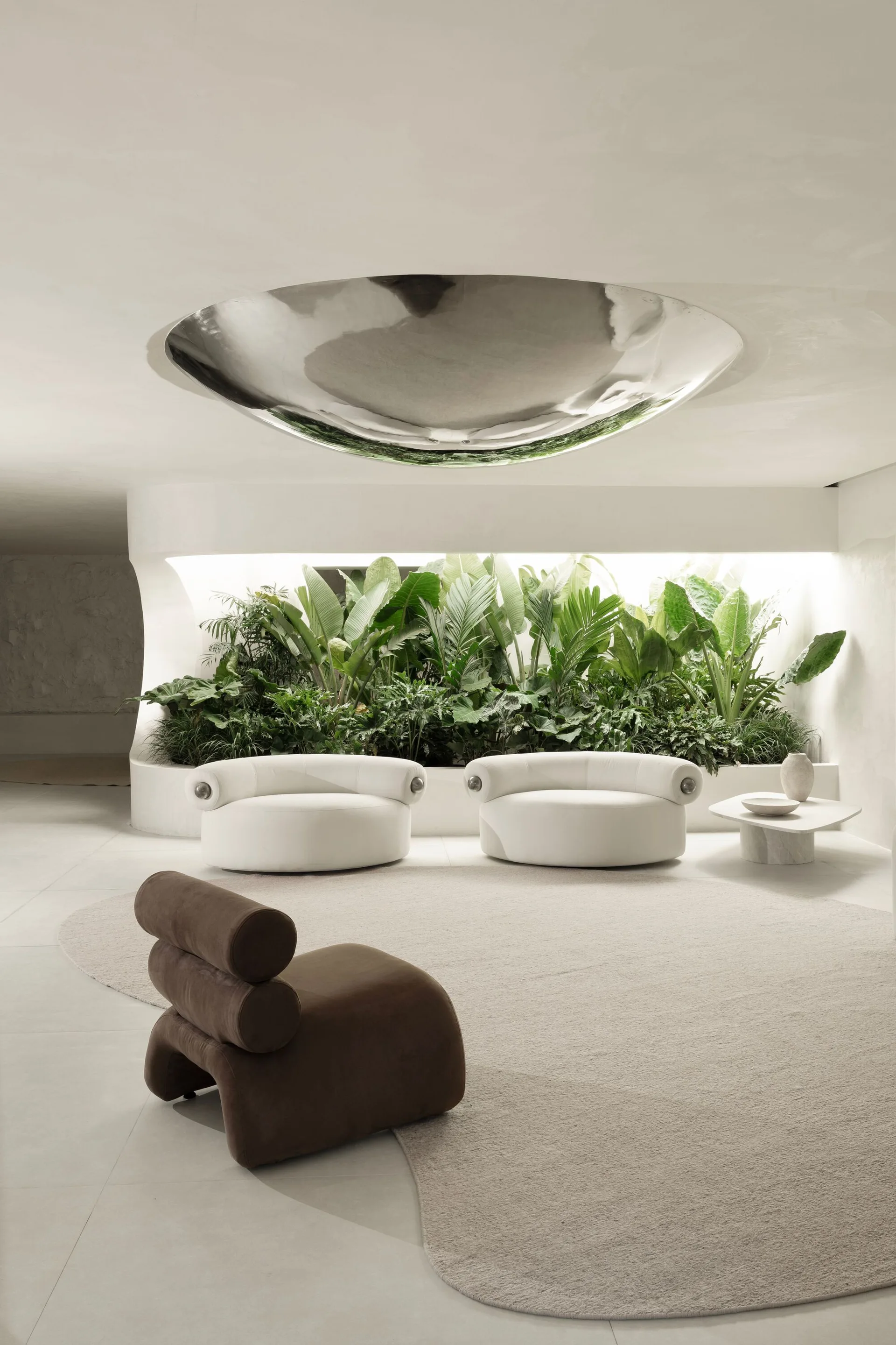A minimalist lounge featuring rounded white armchairs, a brown sculptural chair, and a lush built-in indoor garden bed against curved plaster walls.
