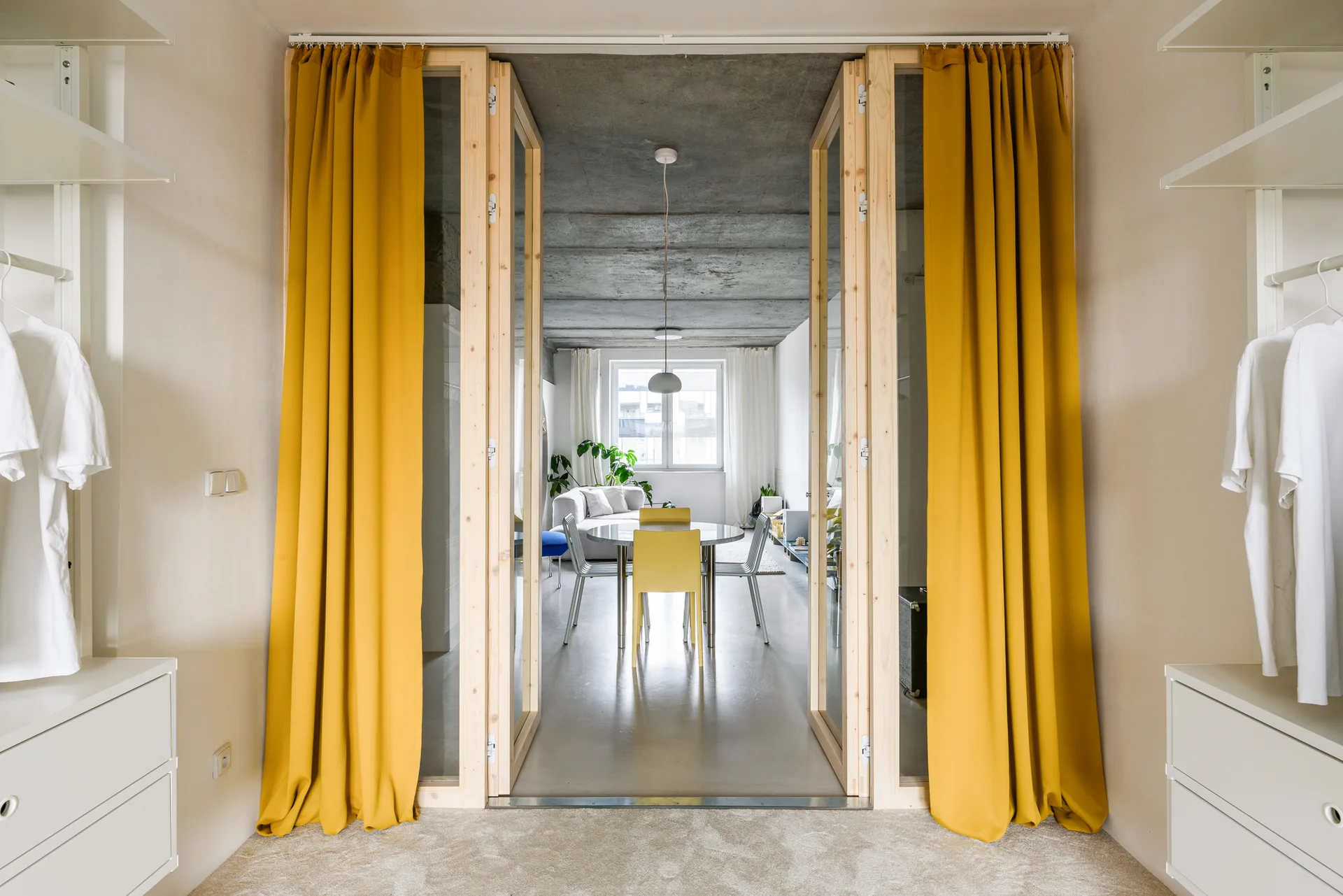 A view through wooden folding glass doors framed by yellow curtains, looking into a modern dining area with a concrete ceiling.