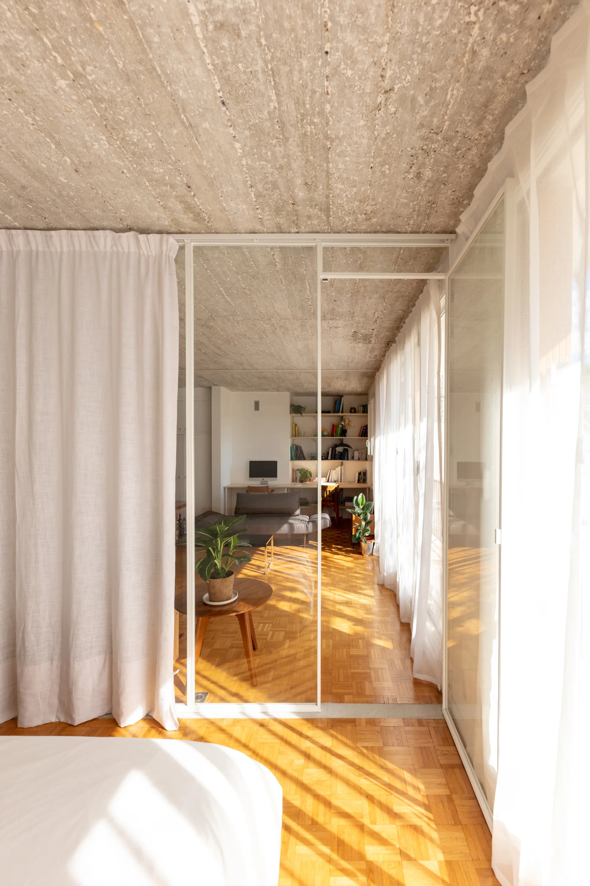 A modern apartment interior features a raw concrete ceiling, a warm wood parquet floor, and a glass partition separating the spaces.