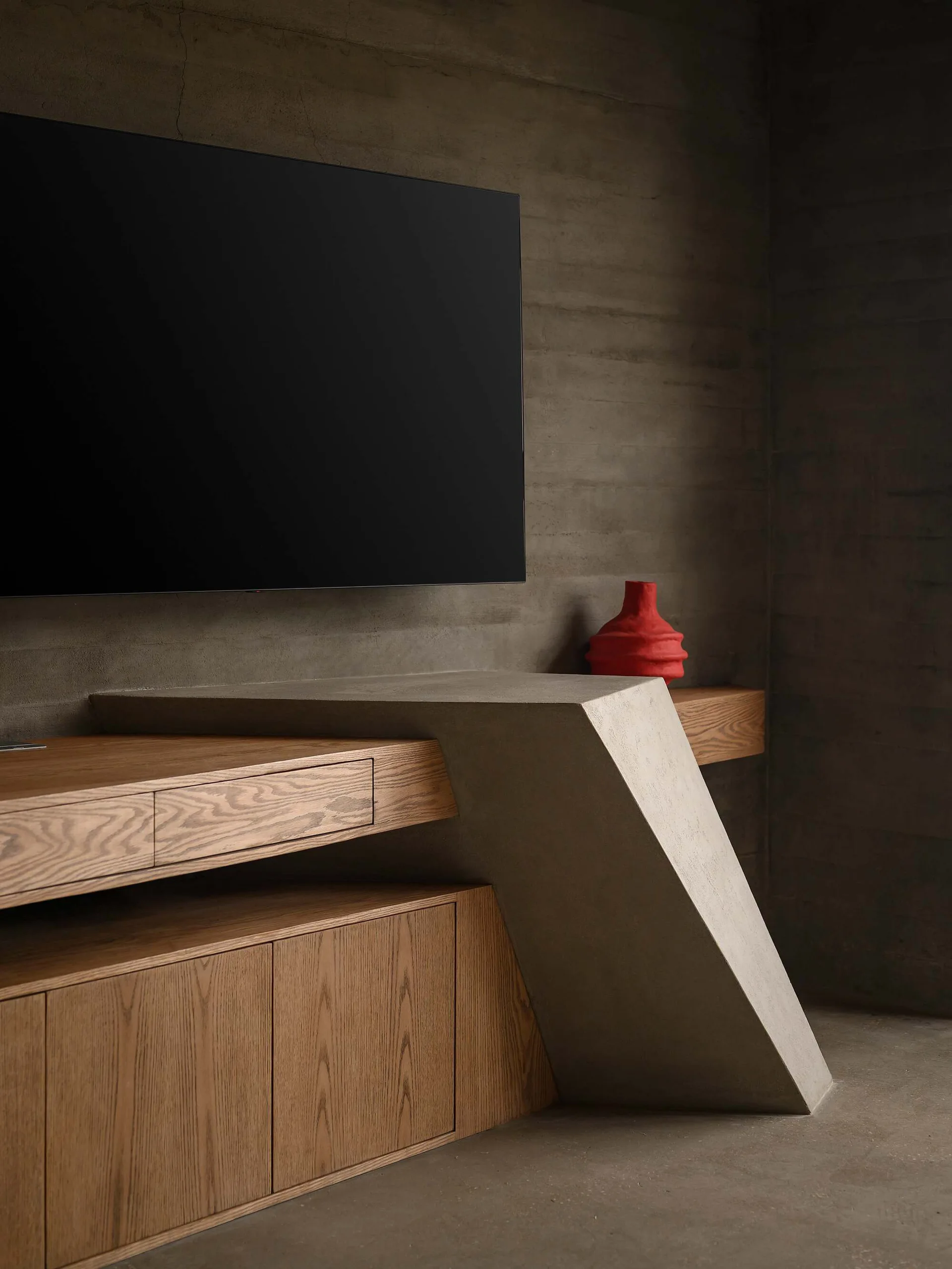 A modern wooden media console features a large angled concrete support column set against a raw concrete wall below a wall-mounted television.
