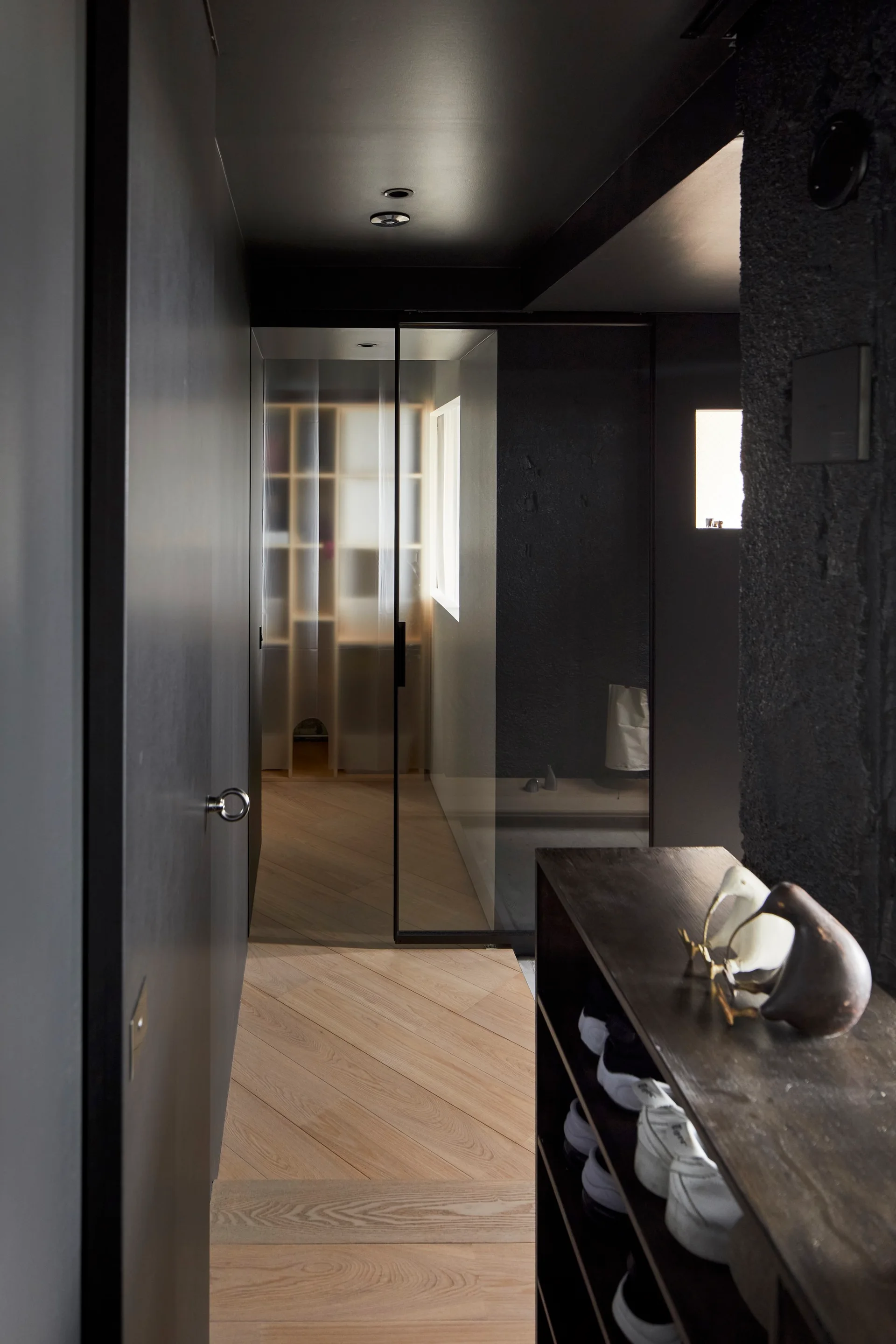 A moody dark gray hallway features diagonal light wood flooring, a black ceiling, and a translucent glass partition at the far end.
