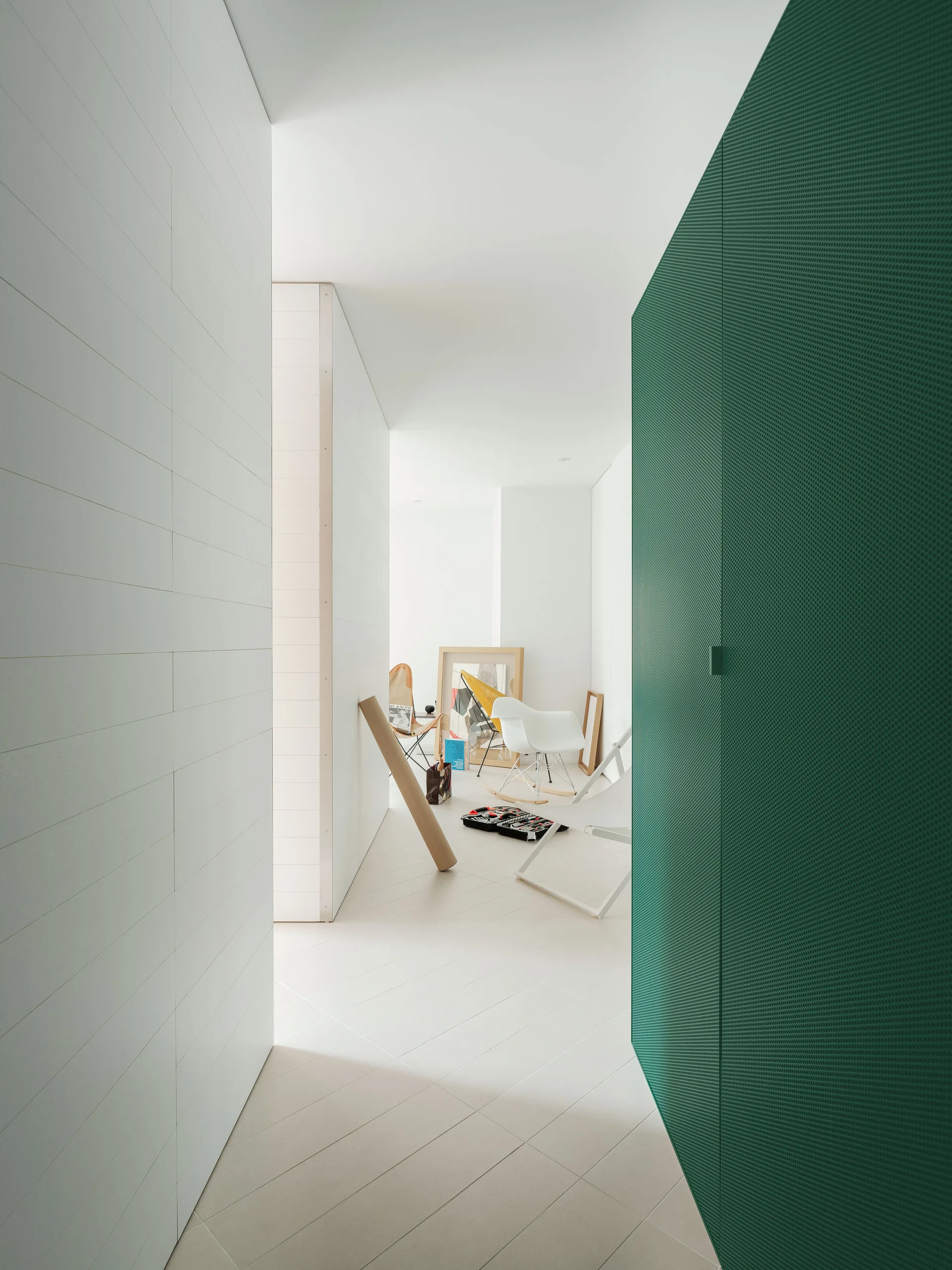 A minimalist white hallway features horizontal wall paneling and a large built-in green perforated metal cabinet leading to a bright back room.