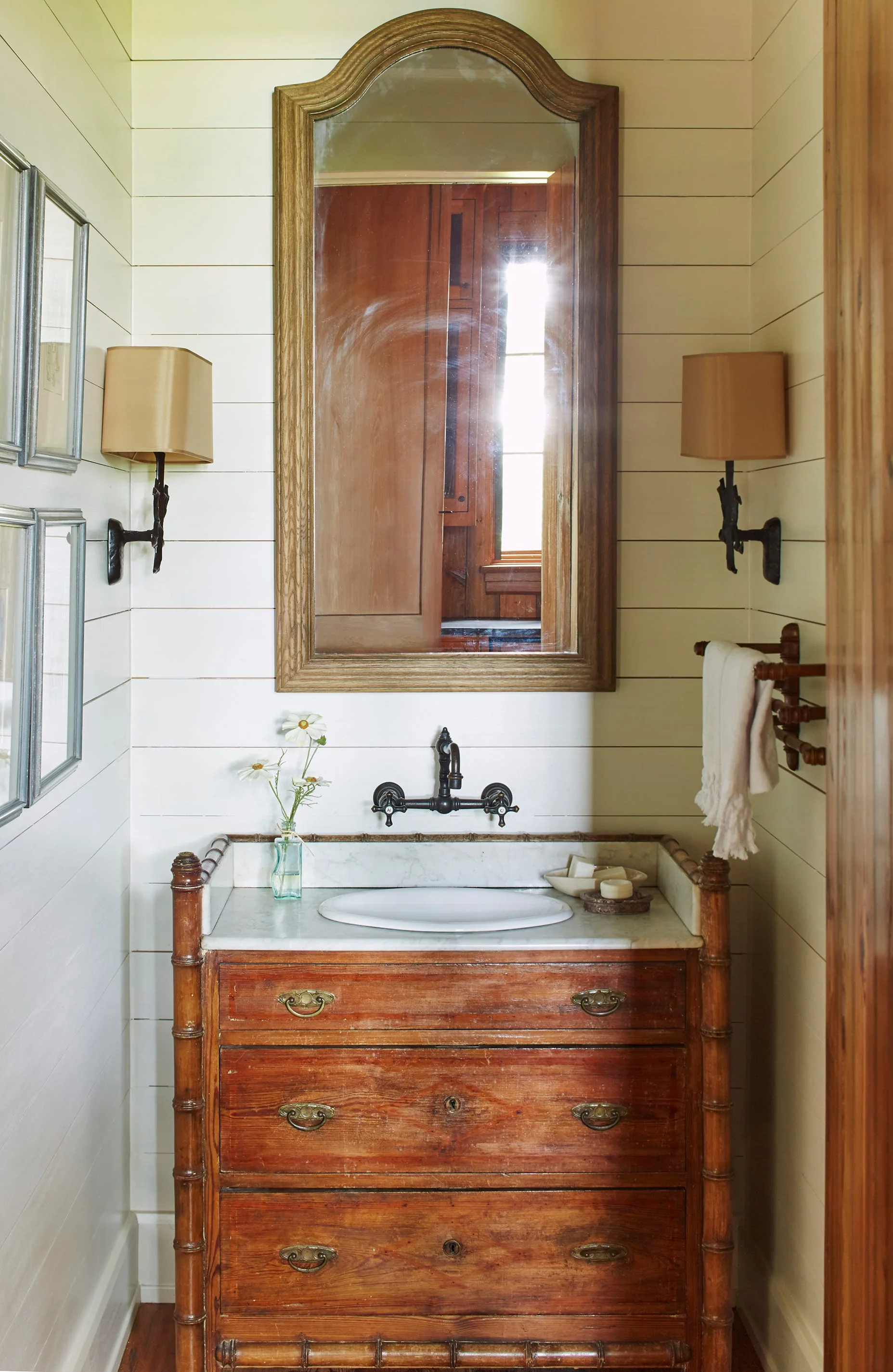 An antique wooden vanity with a marble top sits against a white shiplap wall beneath an arched wooden mirror and two sconces.