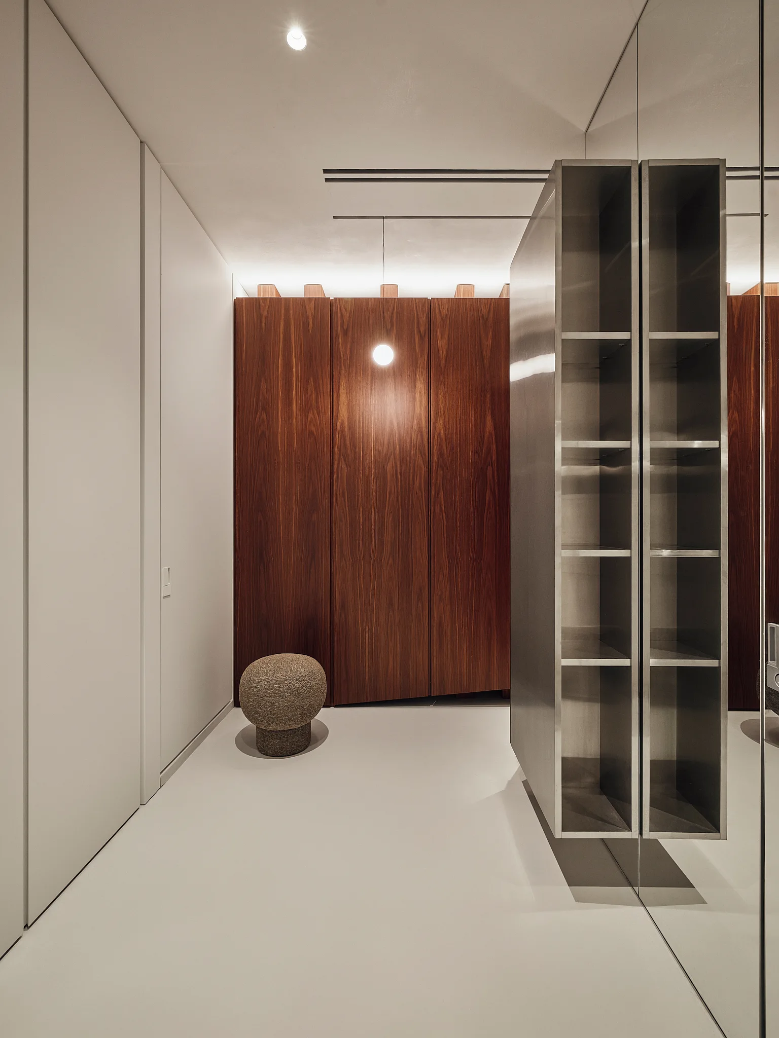 A minimalist dressing area featuring dark wood grain cabinetry, a floating metal shelving unit, a round textured stool, and a large mirror.