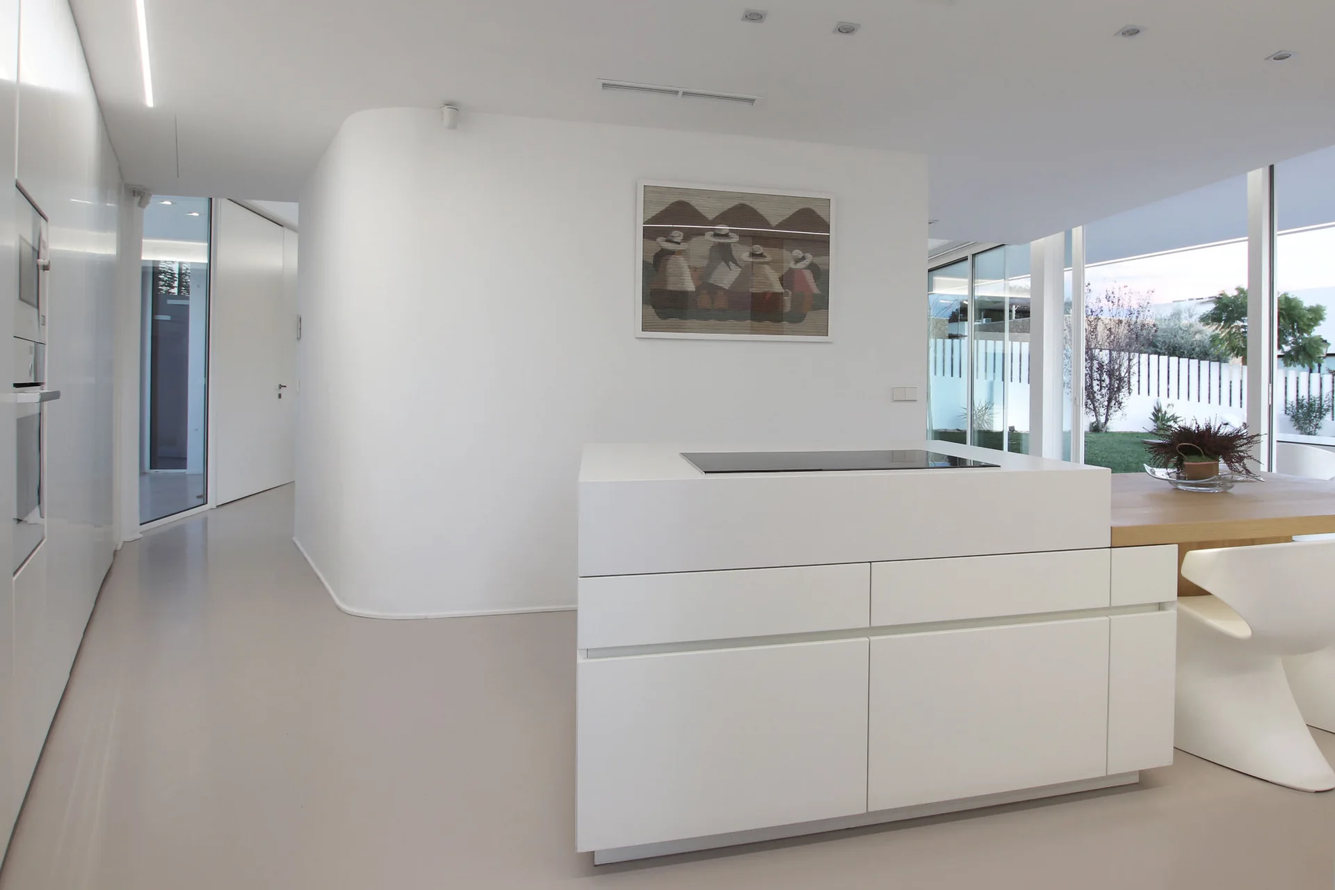 A minimalist white kitchen featuring a central island with a cooktop, a curved partition wall, and a wooden dining table with sculptural chairs.