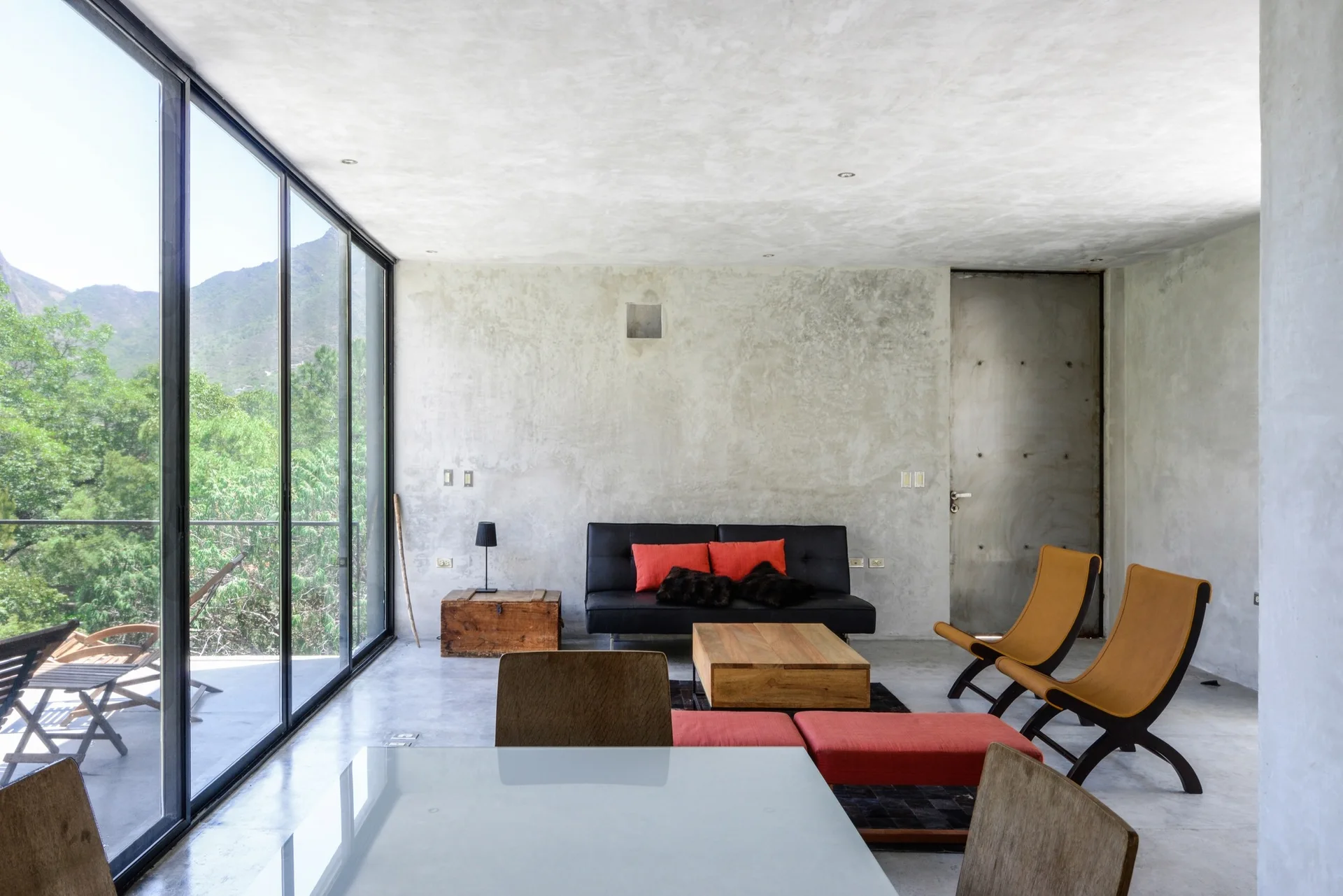 A minimalist living room features exposed concrete walls and floor, a low black sofa with red pillows, tan lounge chairs, and a large glass window wall.