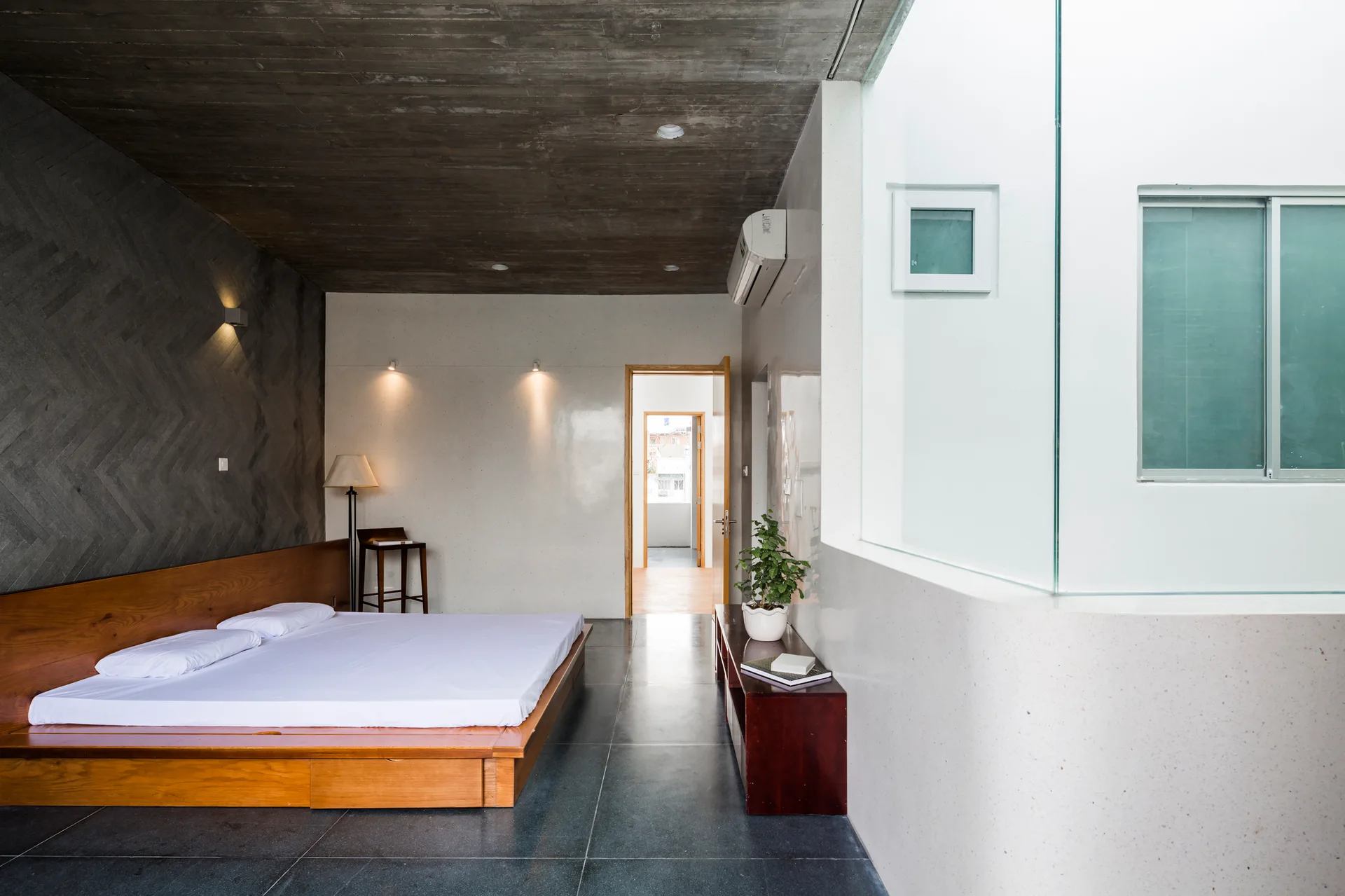 A minimalist bedroom features a wooden platform bed against a grey herringbone stone accent wall, beneath an exposed board-formed concrete ceiling.