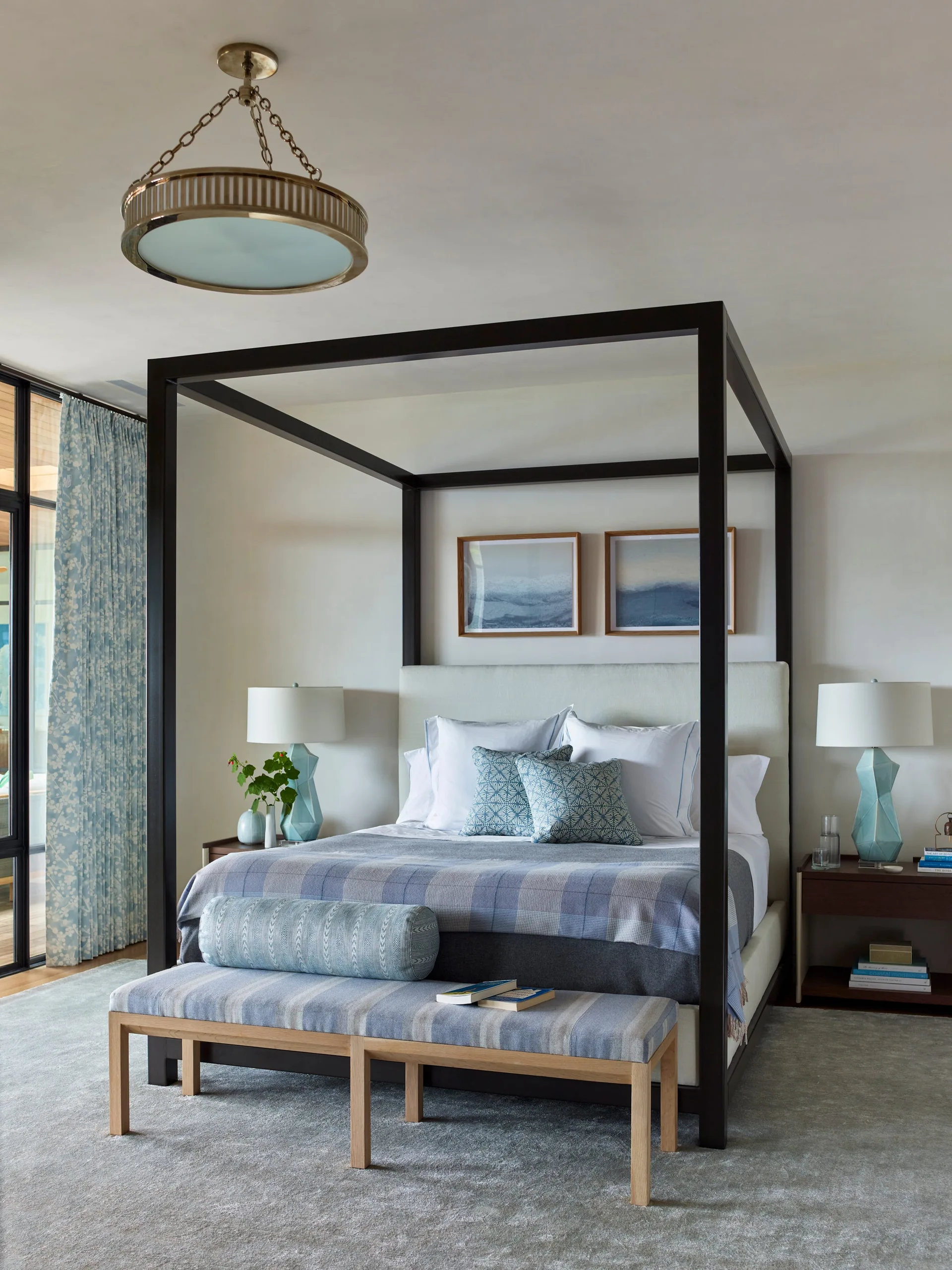 A bedroom features a dark wood canopy bed with blue patterned bedding, a striped bench at the foot, and blue geometric table lamps.