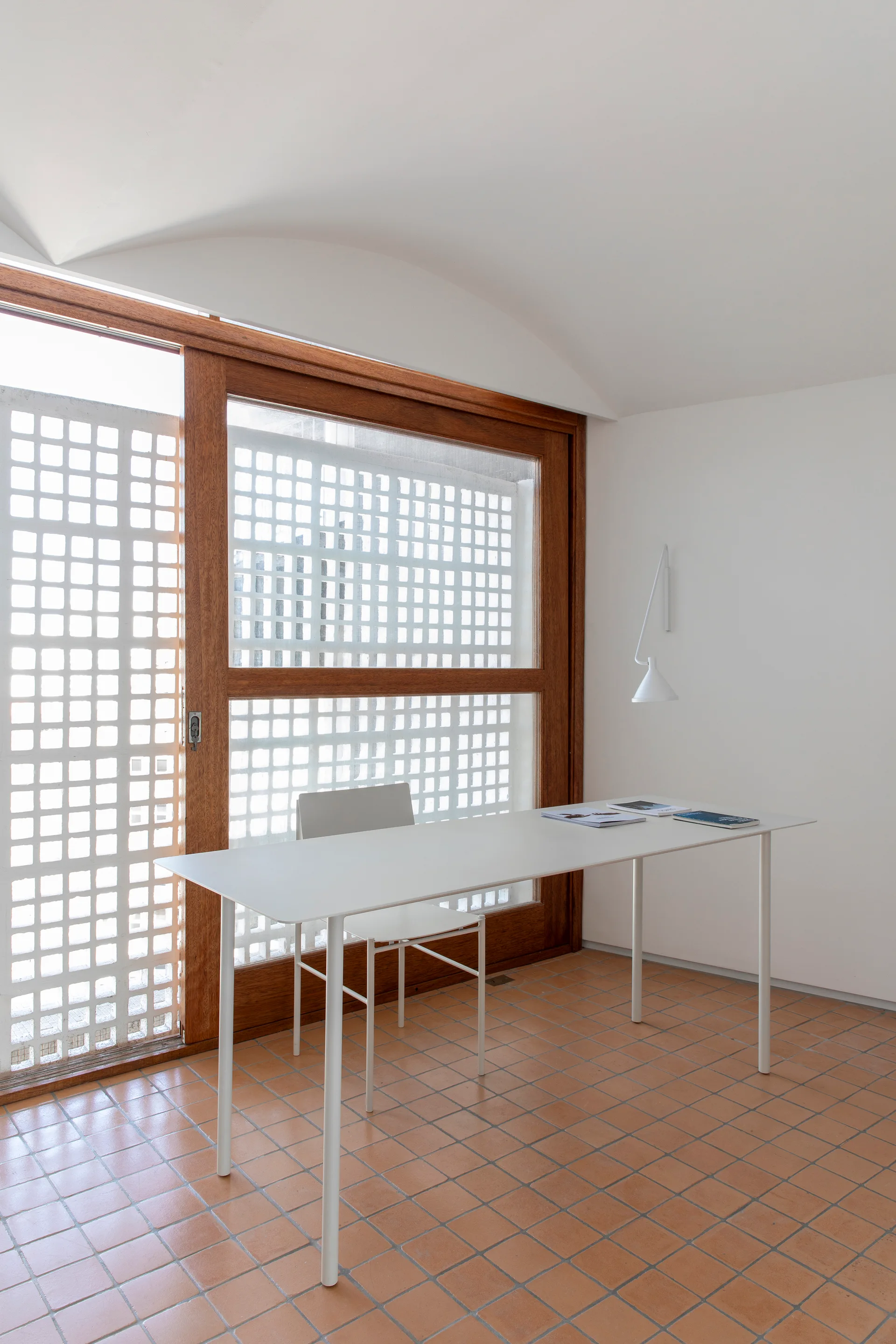 A minimalist white desk and chair sit on terracotta floor tiles in front of a large wood-framed sliding door with a grid-patterned screen.
