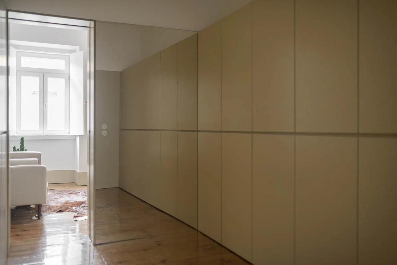 Seamless beige cabinetry lines a hallway floor with dark wood planks, leading to a bright adjacent room with an armchair and shuttered window.