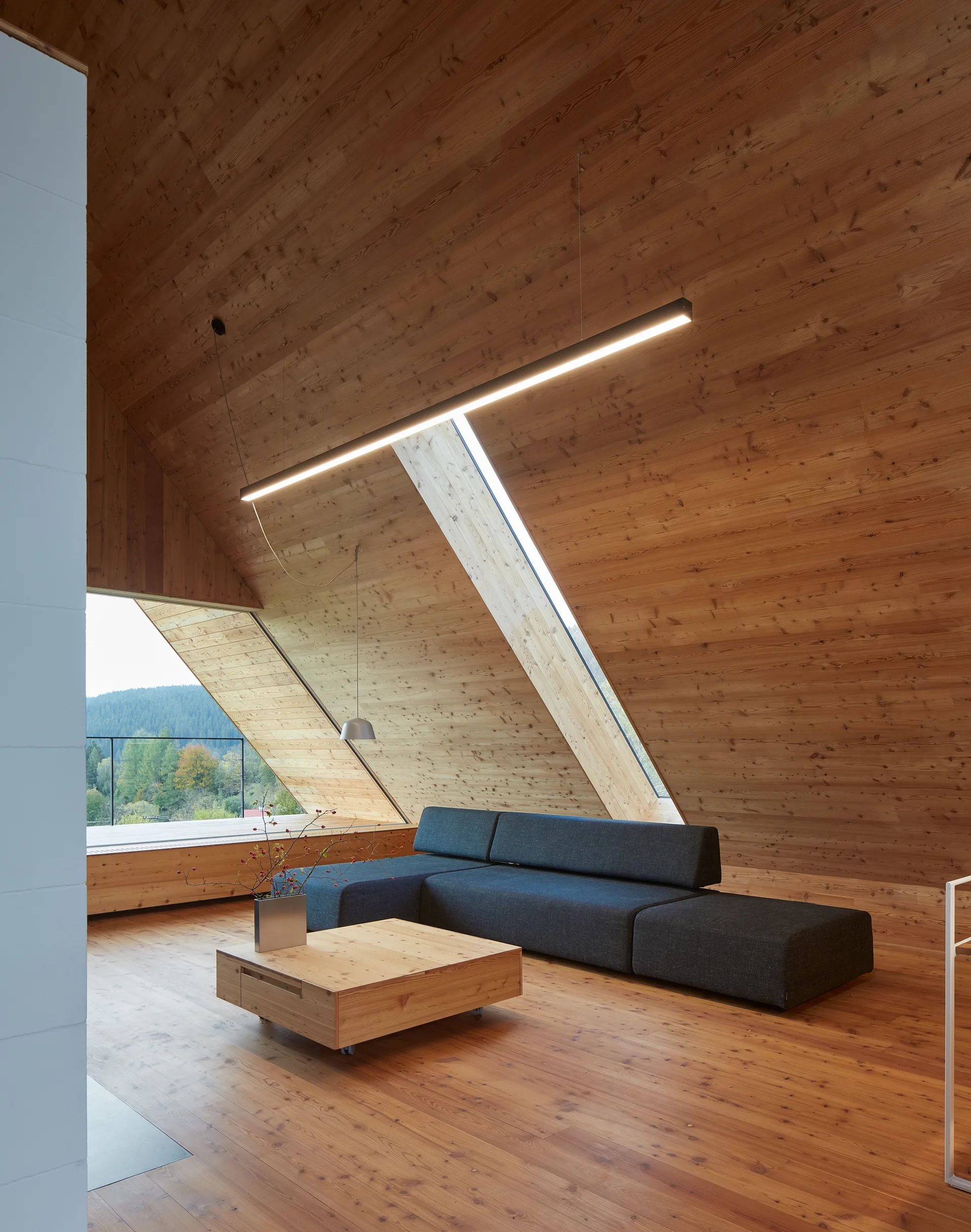 A minimalist attic living room featuring warm wood paneling on the walls and sloped ceiling, a dark grey sectional, and a black linear light.