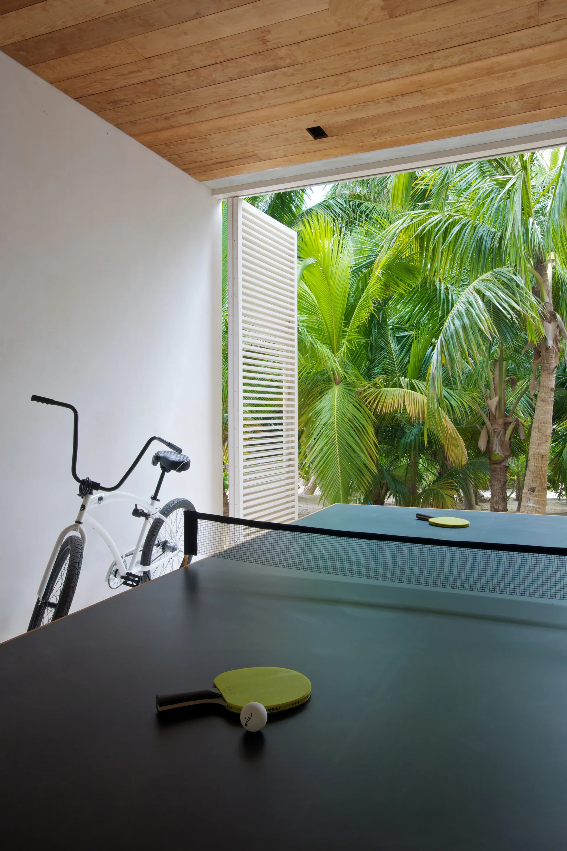 A covered outdoor patio features a black ping pong table, a white bicycle against a plain wall, a wood ceiling, and a view of lush palm trees.