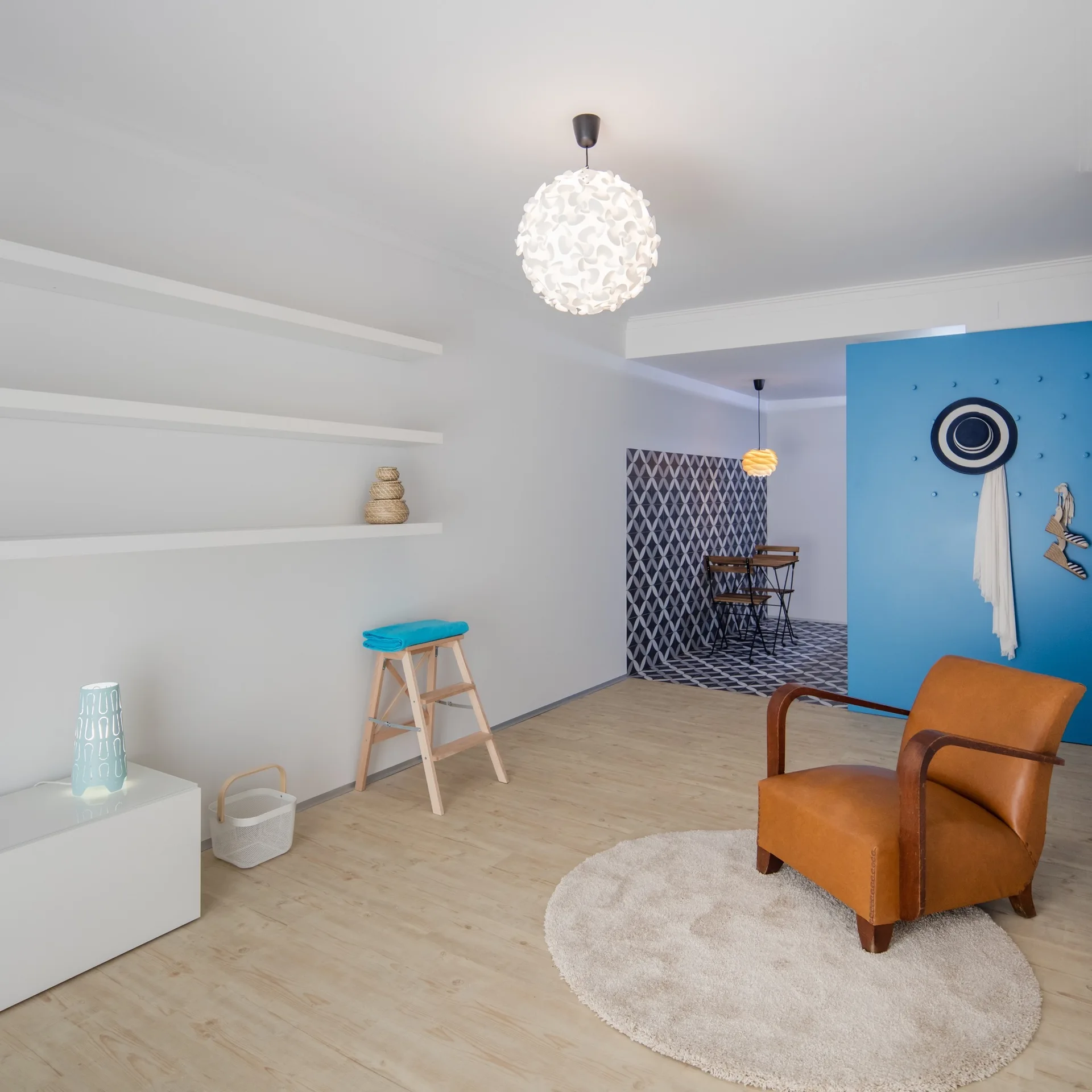 A tan leather armchair sits on a round cream rug in a minimalist living room with white floating shelves and a blue partition wall.
