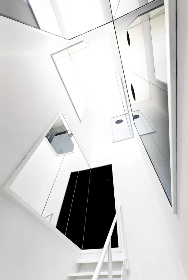 Looking up a minimalist white stairwell with a simple metal handrail and large rectangular mirrors reflecting the surrounding white walls.