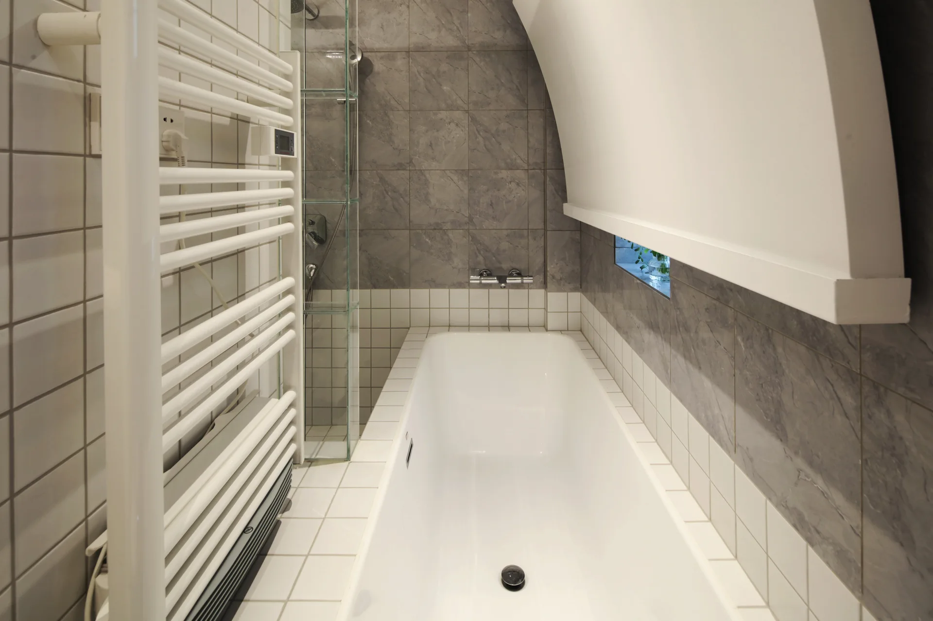 A white inset bathtub is surrounded by white square grid tiles and grey stone-effect tiles, with a tall white ladder towel warmer on the left.