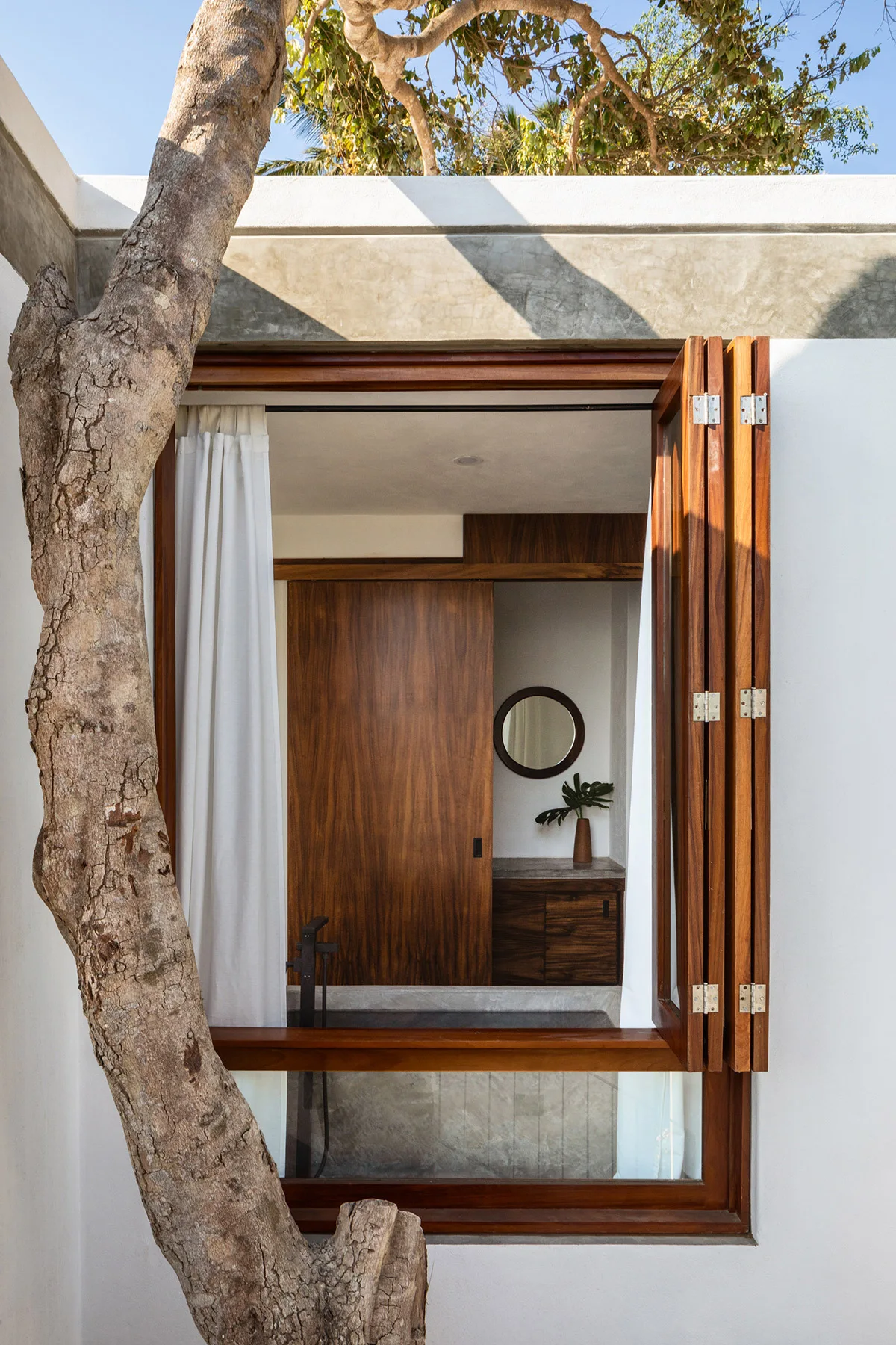 A view through an open wooden bifold window reveals a modern bathroom featuring a dark wood vanity, round mirror, and a white curtain next to a tree trunk.