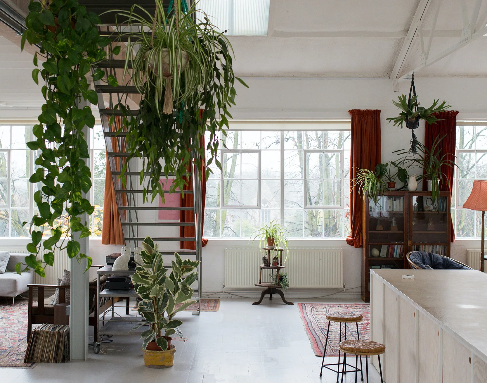 A bright open loft living space featuring a central metal staircase, large industrial windows with orange curtains, and various hanging houseplants.