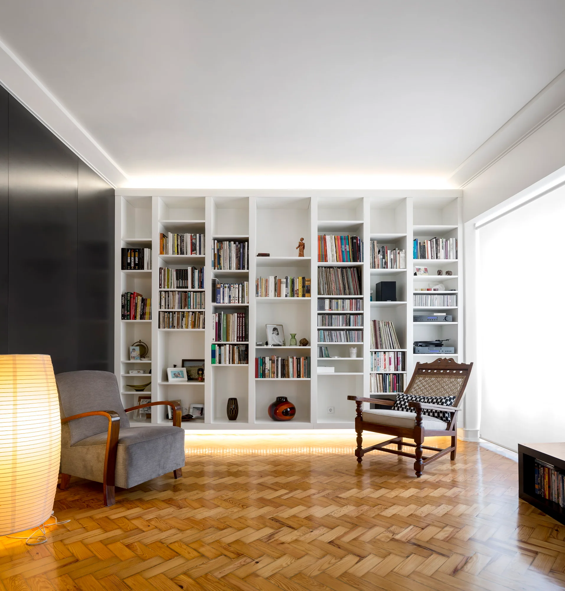 A white built-in bookcase with integrated lighting stands against a wall in a living room featuring herringbone parquet wood floors and two armchairs.
