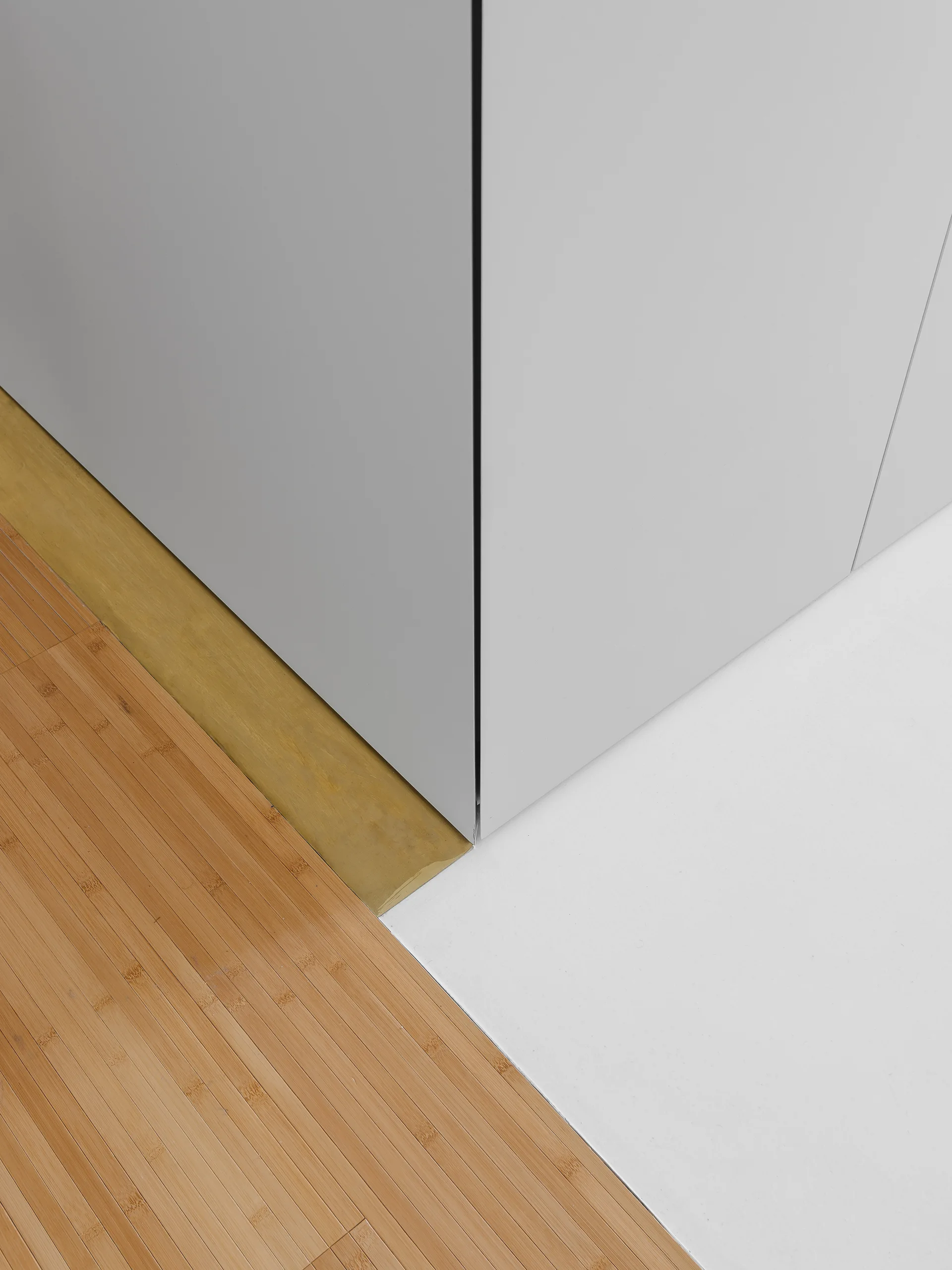 A close-up of a grey cabinet corner where bamboo wood flooring meets a smooth white floor surface with a gold transition strip.