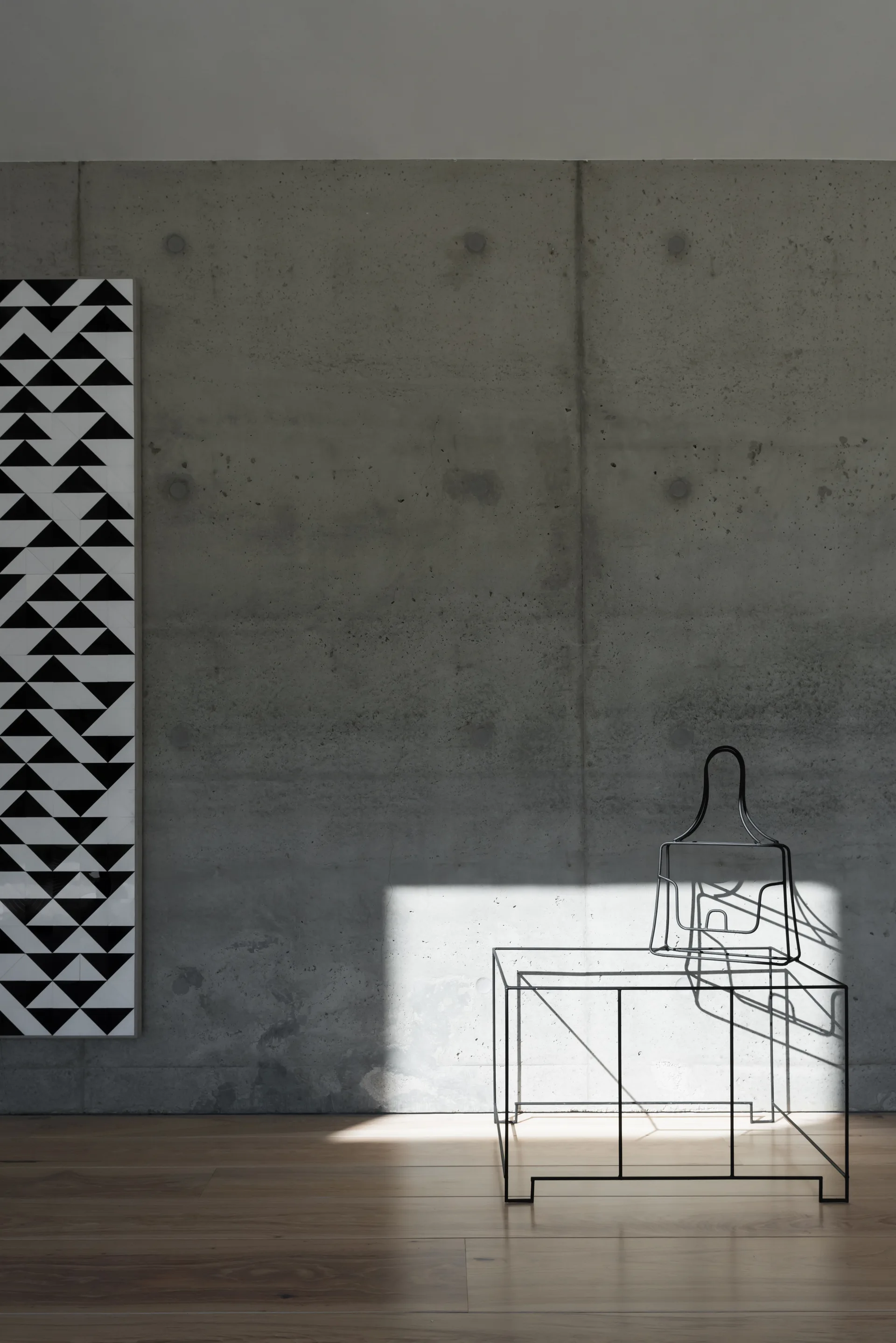 A minimalist room featuring a raw concrete wall, a black wire-frame chair, a black and white geometric art piece, and warm wood floors.