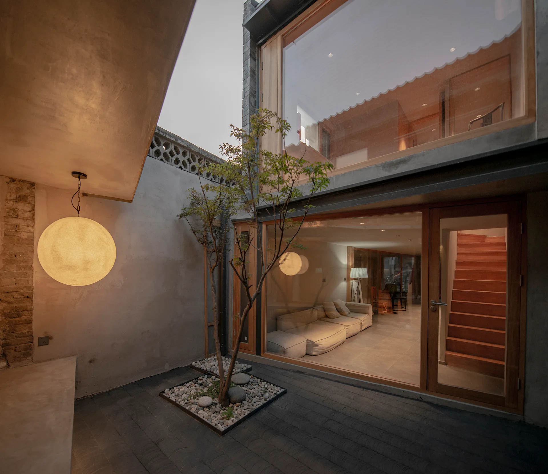 A small concrete courtyard with a tree planted in gravel features a large glass wall looking into a modern wood-trimmed interior.
