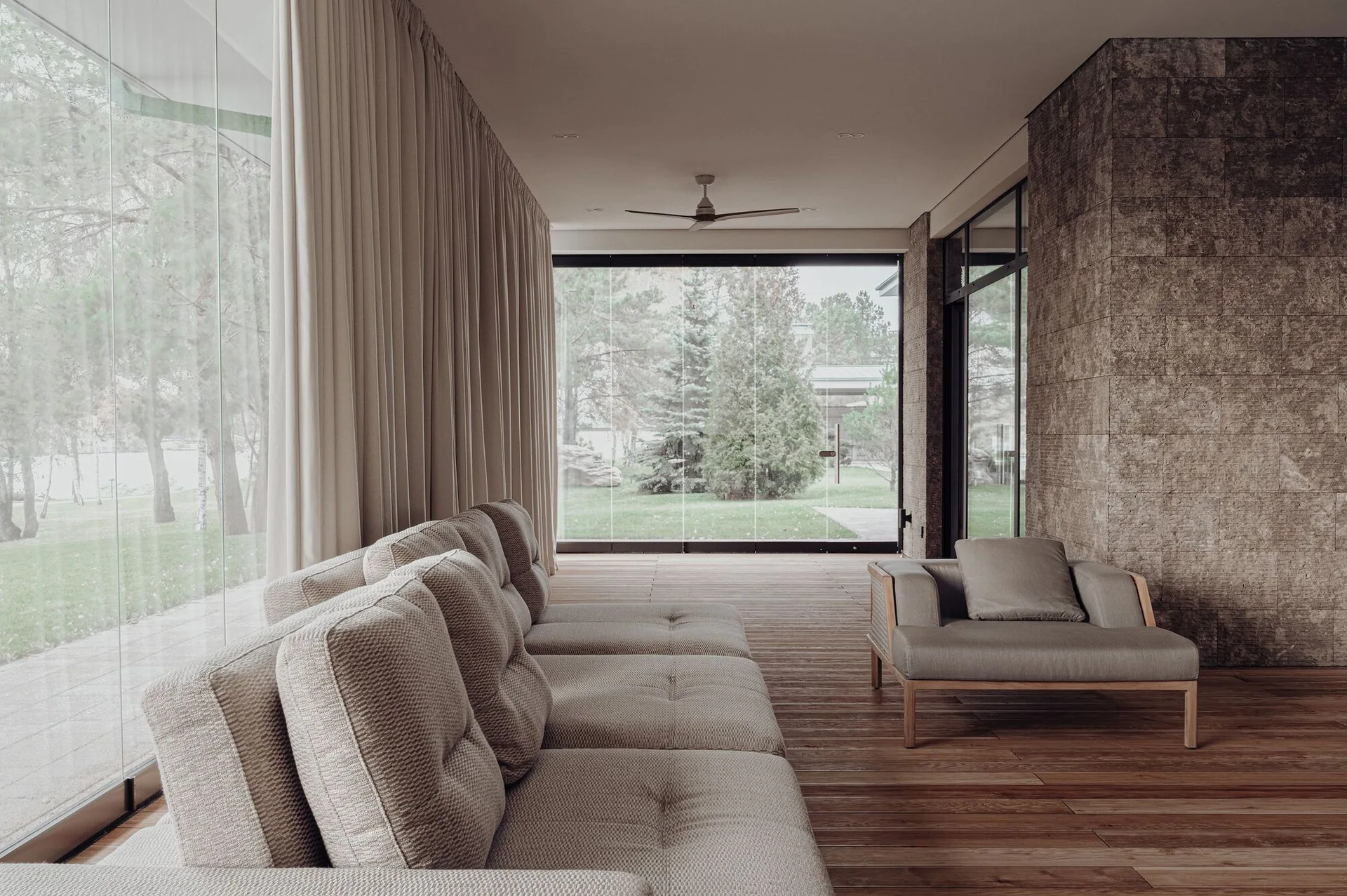 A minimalist living room features a beige sectional sofa, a stone tile accent wall, wood plank floors, and floor-to-ceiling glass walls.