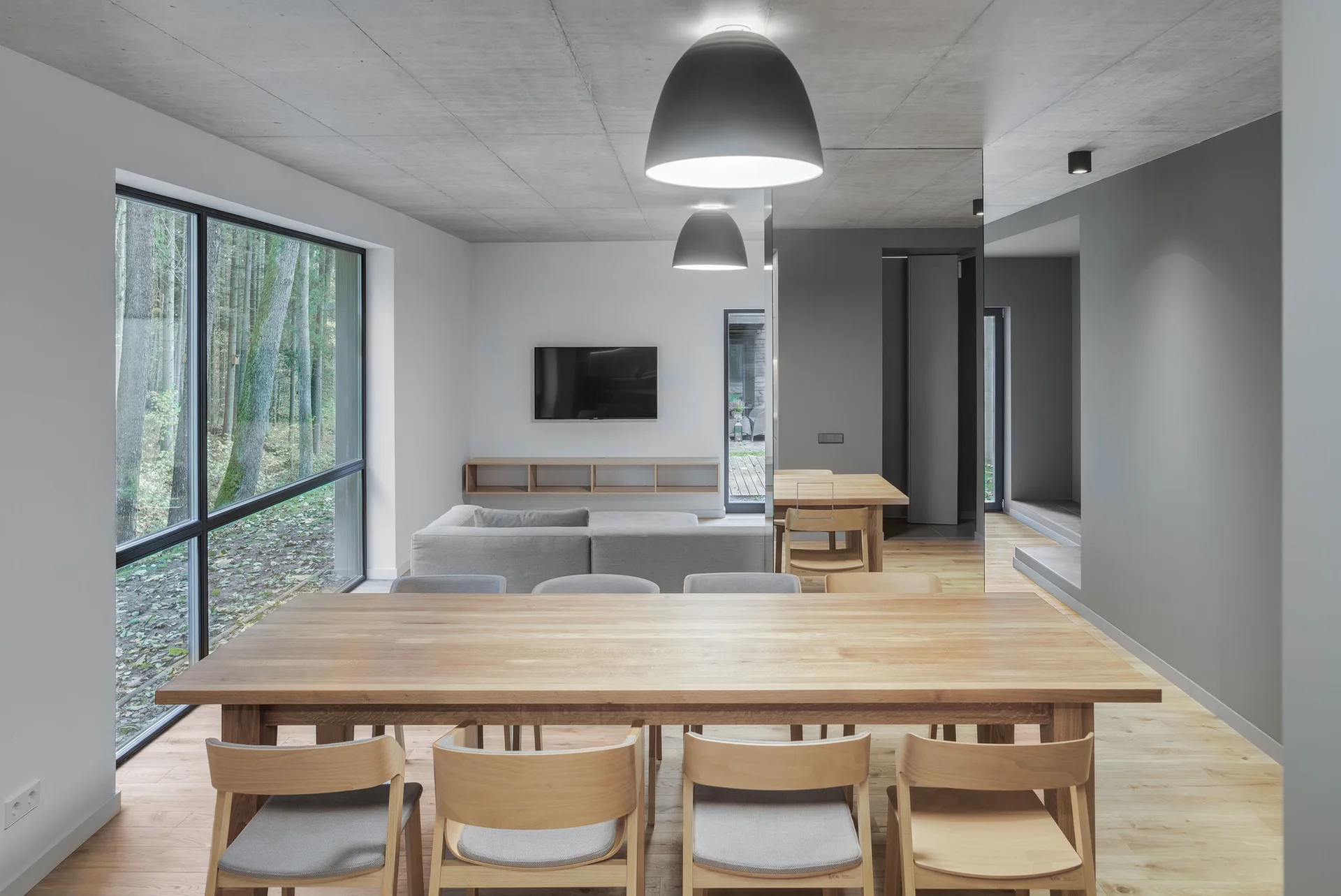 A modern open-plan dining room features a large light wood table, wooden chairs with grey cushions, a concrete ceiling, and large dome pendant lights.
